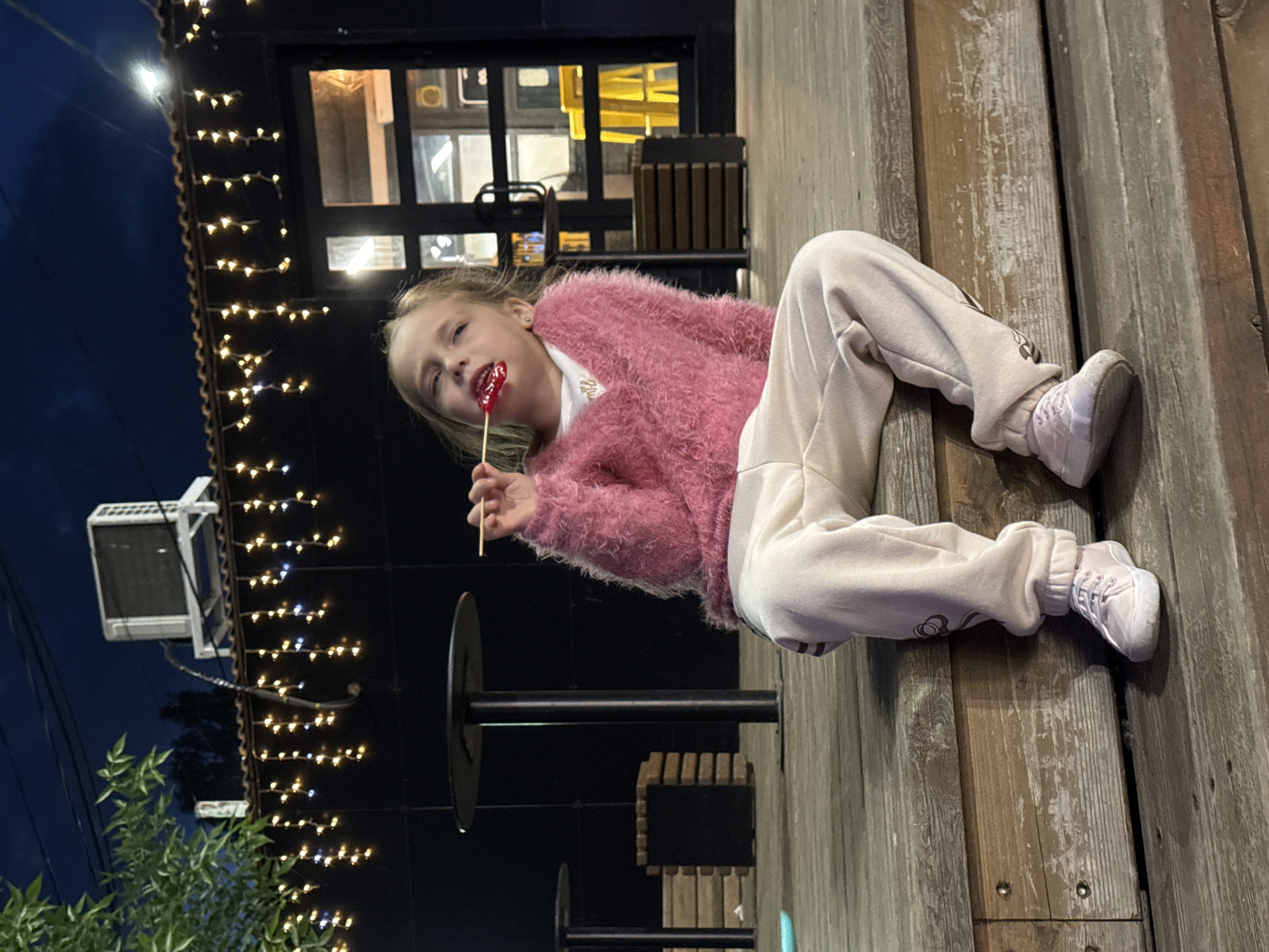 A young girl with blonde hair is depicted lying on her left side on a wooden surface, possibly a deck or outdoor platform, in Izhevsk, Russia. She is wearing a fluffy pink sweater over a white top, light-colored sweatpants, and light sneakers. In her right hand, she holds a red lollipop on a stick, which she is putting into her mouth, with some of the red color visible on her lips. She looks directly towards the camera with a slight, playful expression.
The setting appears to be evening or night, indicated by the dark sky in the background and numerous warm white string lights adorning the eaves and facade of a building. The building features large, dark-framed windows, suggesting a cafe, restaurant, or similar establishment. Behind the girl, various elements of the outdoor area are visible, including what looks like dark metal tables or stools. In the upper left corner, an air conditioning unit is mounted on a wall, and some green foliage is visible at the bottom left. The wooden surface she is on shows signs of wear. No discernible text is visible in the image.