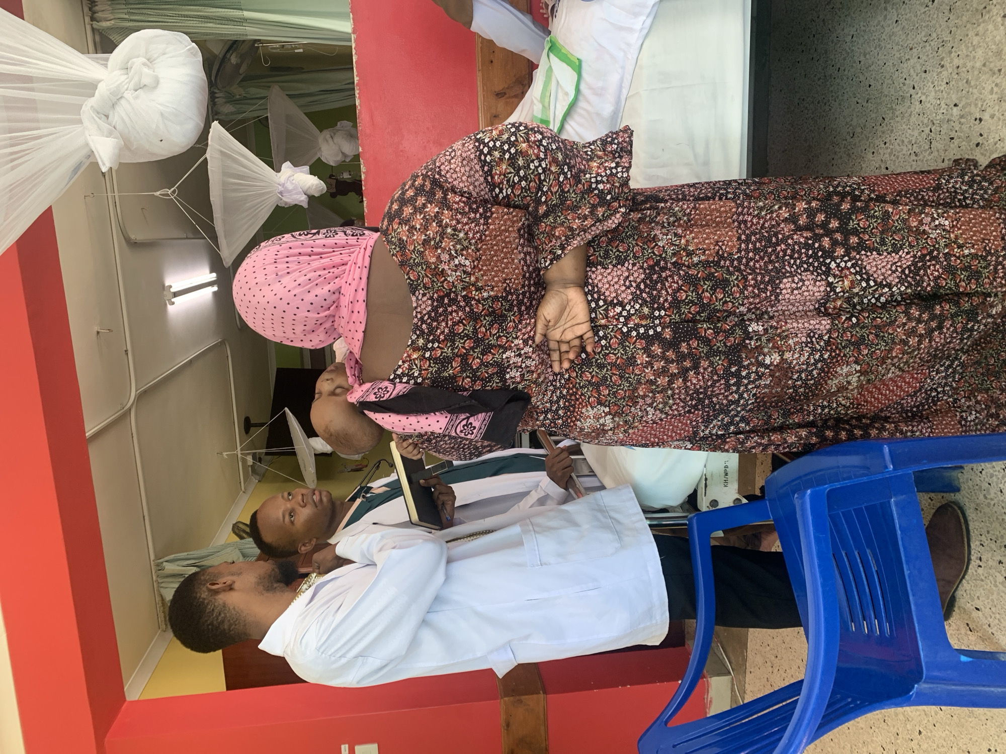 A woman in a floral dress and pink headscarf stands next to a hospital bed, talking to a man in a white coat who holds a notebook. Another man in a white coat stands to the side, listening. The setting appears to be a hospital room with mosquito nets hanging from the ceiling. A blue plastic chair is also visible. The location is likely Dar es-Salaam, Tanzania, given the mosquito nets, and the appearance of the people in the image.