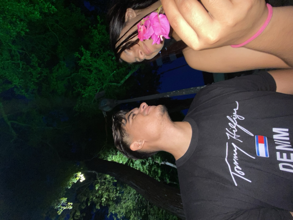 A nighttime outdoor scene shows a man looking up at a woman. She holds a pink flower in front of her face. The man wears a black Tommy Hilfiger t-shirt. Dark trees fill the left of the frame. The overall composition is shot from a low angle. The subjects appear to be enjoying a quiet moment together, illuminated by a soft light source.