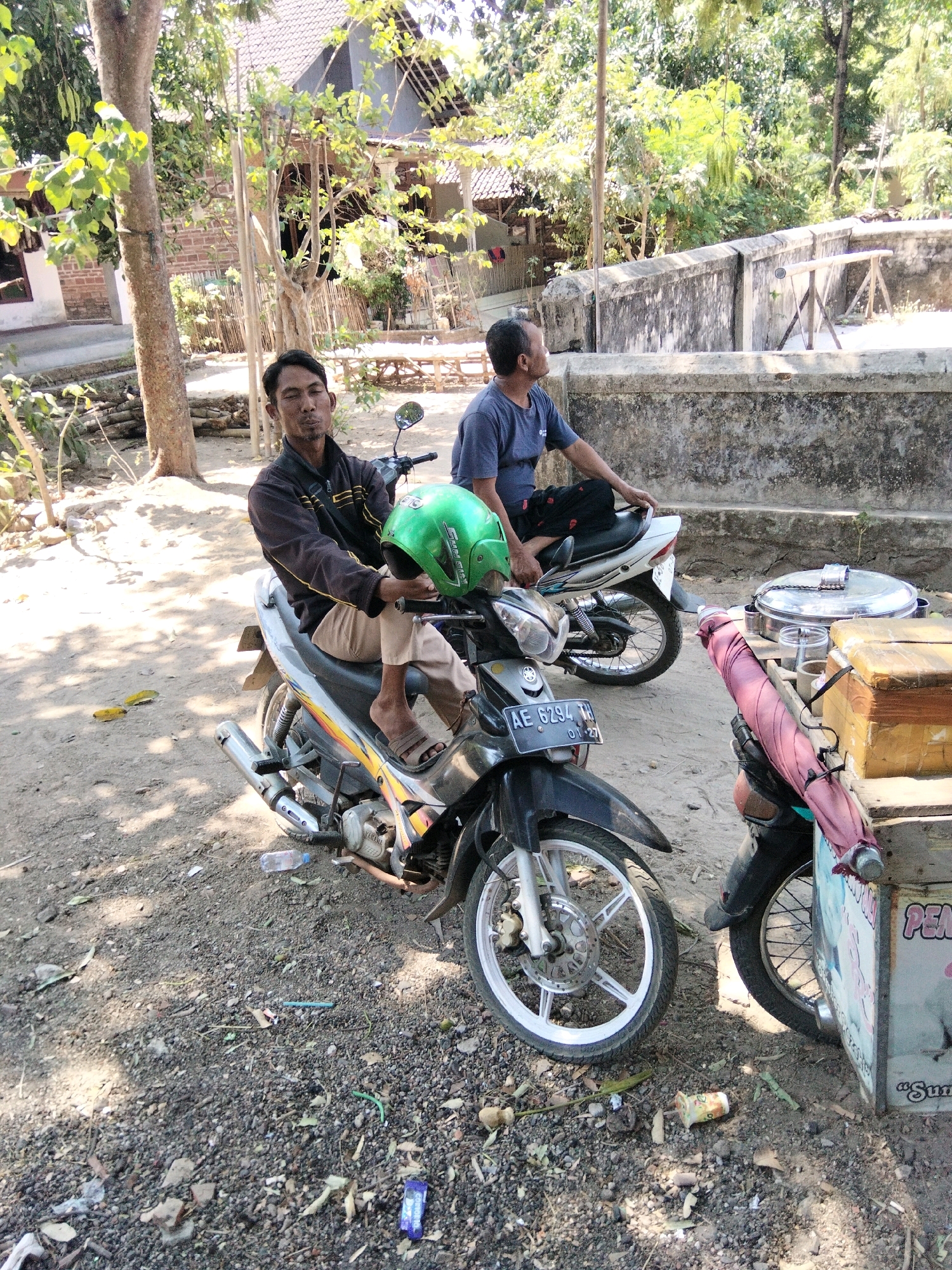 The image depicts an outdoor scene in Jenangan, Indonesia, likely a village or rural area, characterized by dusty ground, sparse vegetation, and simple buildings. In the foreground, two men are seated on motorbikes. The man on the left, wearing a black jacket and tan pants, holds a green helmet in his left hand. He is looking directly at the camera with a neutral expression. His motorbike is black and silver with visible engine details and a license plate reading "AE 6294 TH". To his right, a second man, wearing a blue t-shirt, is seated on a white motorbike, looking away from the camera towards the right. His motorbike is partially obscured. Between the motorbikes, a wooden cart with a pink umbrella and various containers is visible. The cart appears to be used for vending. In the background, there are trees, a low wall, and a building with a tiled roof, suggesting a residential or community area. The lighting indicates it is daytime, with sunlight casting shadows. The ground is covered in gravel, dirt, and scattered debris.