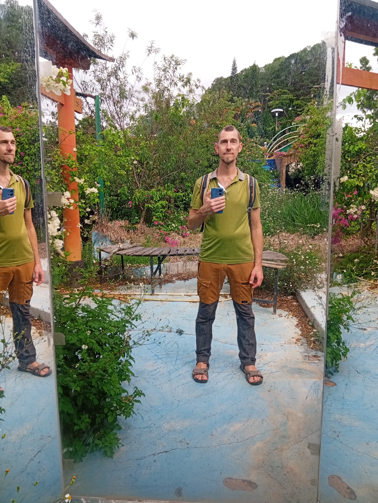 A man stands in front of a large, slightly dirty mirror, in an outdoor area with a blue cracked surface, resembling a patio or walkway. Behind the man is a garden with diverse greenery, trees, and a wooden bench. The man has short brown hair, a beard, and wears a green polo shirt, brown cargo pants, and sandals. He's holding a blue smartphone. The reflection in the mirror shows an identical scene, mirroring the man and the surrounding environment. An orange decorative architectural feature, possibly part of a gazebo, is visible behind the man, along with other garden structures. The location is Da Lat, Vietnam, recognized by its characteristic landscapes, vegetation, and architectural styles.