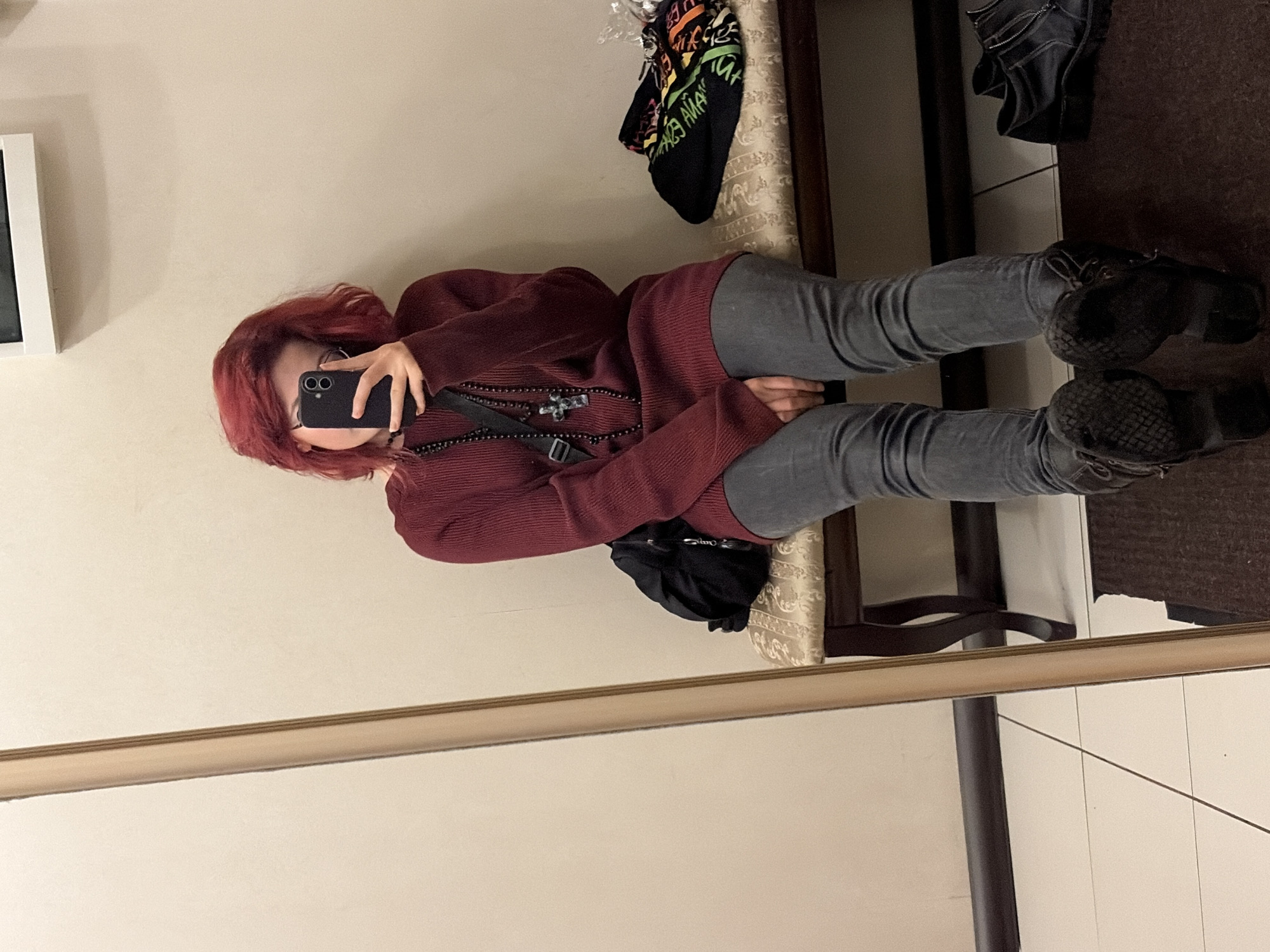 In an indoor setting, a person with short, red hair sits on a bench, taking a mirror selfie with a smartphone. They are wearing a maroon sweater, gray jeans, and dark boots. A black bag is on the bench next to them, and a folded garment with colorful lettering lies on the far end of the bench. A large mirror is positioned to the left of the frame. The floor is tiled. The environment appears to be a dressing room or a public waiting area. There is no indication of the specific location in Chelyabinsk, Russia.