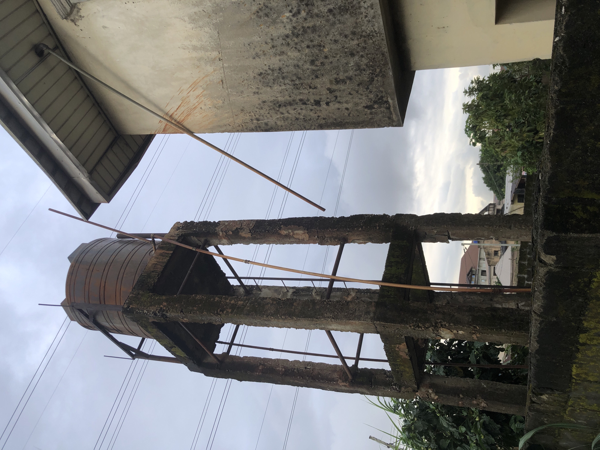 A rusted, cylindrical water tank sits atop a dilapidated concrete structure with four pillars. The structure appears weathered and aged. To the right, a light-colored building with a grey, corrugated roof juts into the frame. A metal pipe extends from this building at an angle, possibly a support or conduit. Power lines are visible across the cloudy sky, crisscrossing above the water tower. In the background, a few other buildings and lush green trees are discernible. The scene is set against a backdrop of overcast skies. The location is in the city of Ado, Nigeria.