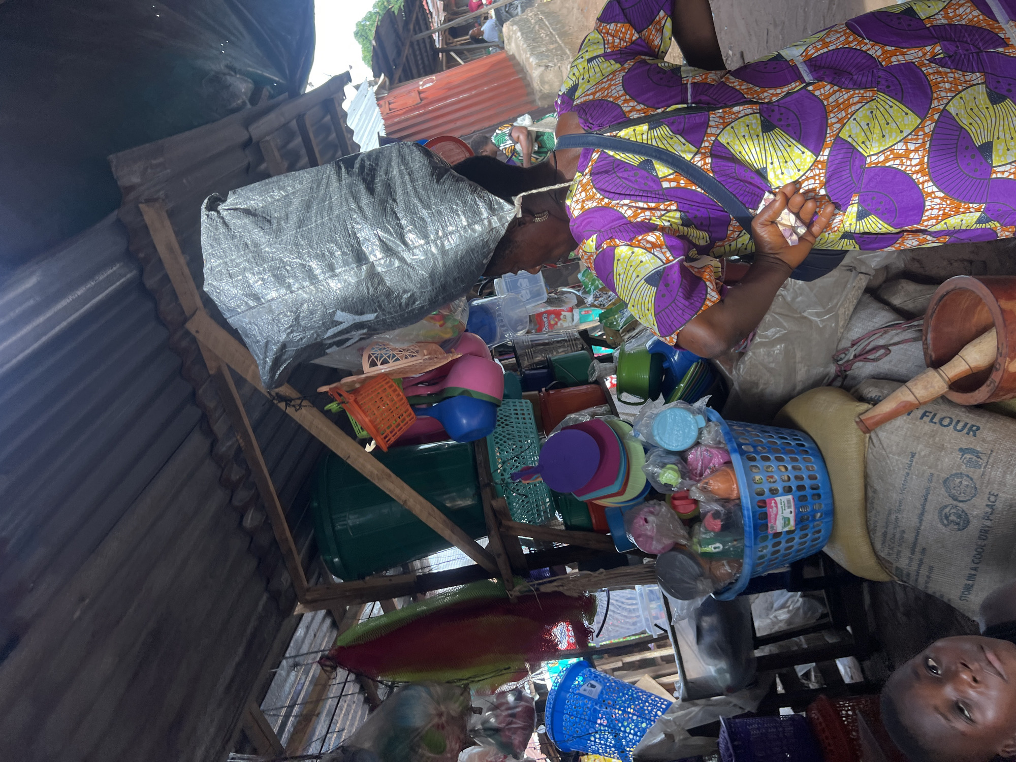 The image captures a bustling market scene in an unspecified location within the city of Unknown, Unknown. The foreground features a person in vibrant purple and yellow patterned clothing handling money, while surrounded by an assortment of goods including plastic bowls, buckets, baskets, and a bag labeled "FLOUR." A second person is partially visible at the bottom of the frame. The market is under a corrugated iron roof, with goods stacked high on wooden structures. A large gray sack dominates the upper left, obscuring the background.
