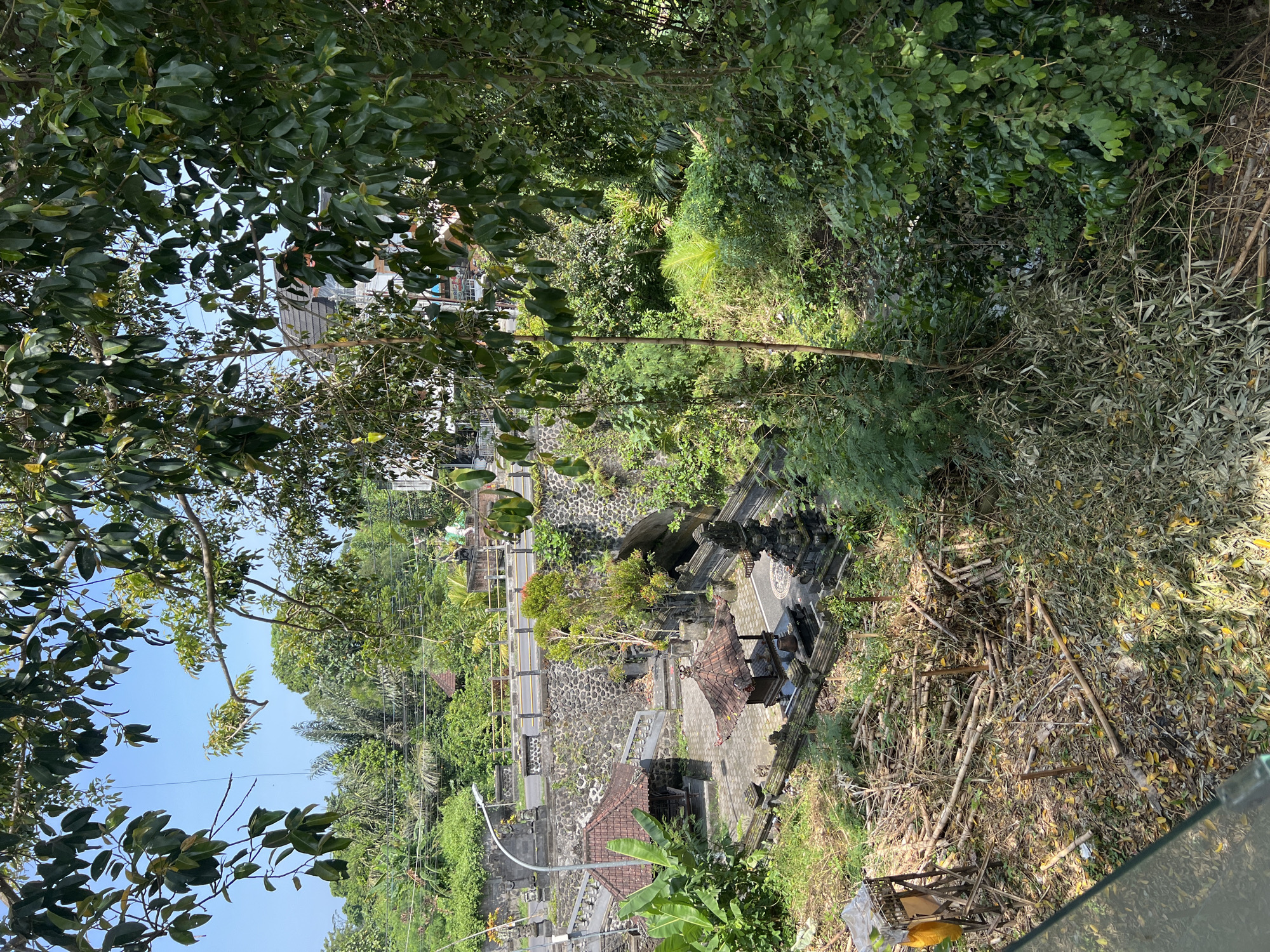 The image shows an elevated view of a temple complex nestled within lush greenery in Buduk, Indonesia. The temple features stone structures, a tiled roof building, and decorative statues. A road with a guardrail runs alongside the temple, suggesting accessibility. In the foreground, there's a cluttered area with bamboo poles, leaves, and some debris, indicating a recent clearing or natural decay. The surrounding environment is densely vegetated with trees and plants, typical of a tropical climate. The sky is visible through the trees.