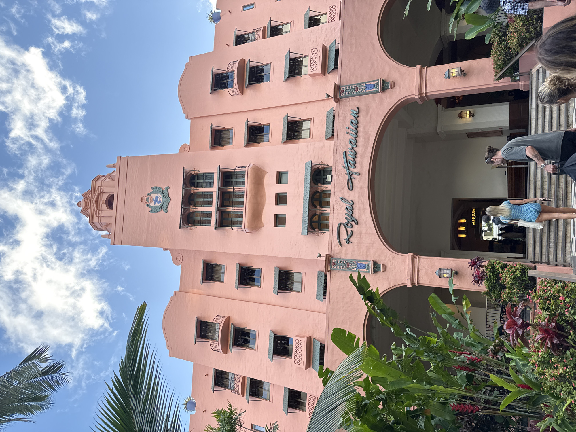 The image depicts the Royal Hawaiian Hotel, a historic landmark located in Honolulu, United States. The hotel is prominently featured with its distinctive pink facade, multiple stories, windows with shutters, and a central tower topped with decorative elements. The "Royal Hawaiian" sign is visible above the hotel's entrance, framed by an archway. People are seen entering and exiting the hotel, and lush tropical plants and greenery frame the lower portion of the image. The sky is blue with scattered clouds.
