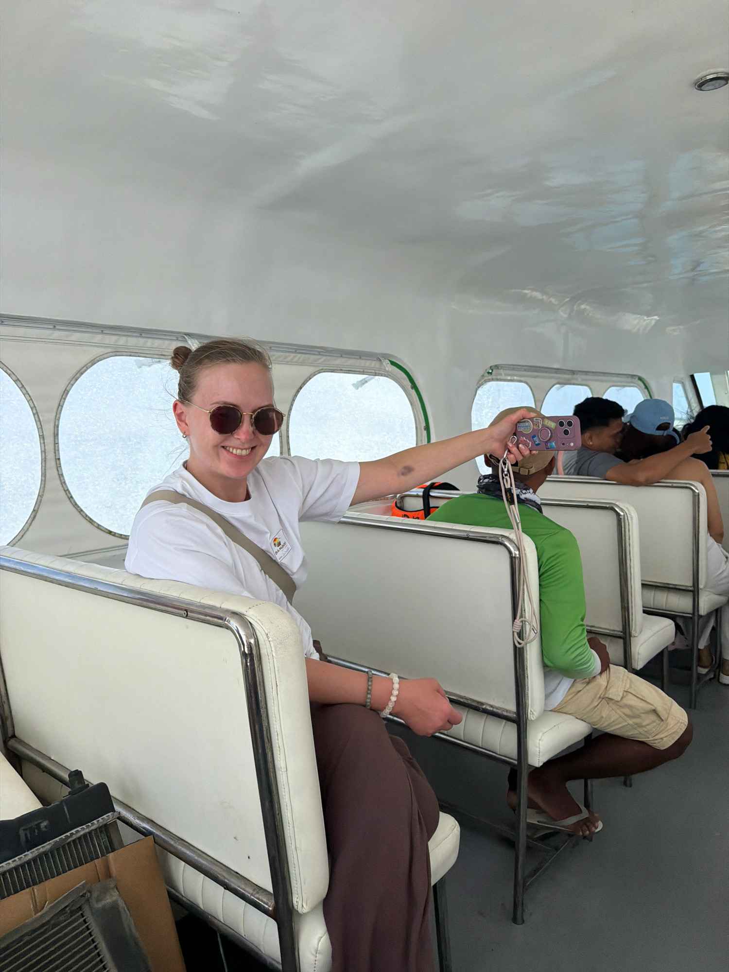 A woman with blonde hair pulled back in a bun, wearing sunglasses and a white t-shirt, is seated on a white bench inside a boat. She is smiling and holding up a phone, seemingly taking a selfie or a photo of the people behind her. The boat has rows of white benches with metal frames and large circular windows on the side. Several other passengers are visible, some seated and others appearing to be in conversation. In the background, there are hints of a city or coastal environment seen through the windows. The woman is wearing a lanyard around her neck, which is attached to her phone. The boat interior appears to be clean and well-lit, with a white ceiling and grey flooring. There are also some storage items visible near the woman, including what appears to be a grille and a cardboard box. The overall impression is a casual and relaxed atmosphere on a boat trip. The location, Malay, Philippines, is known for its beautiful islands and beaches, suggesting this could be a ferry or boat tour in the area.