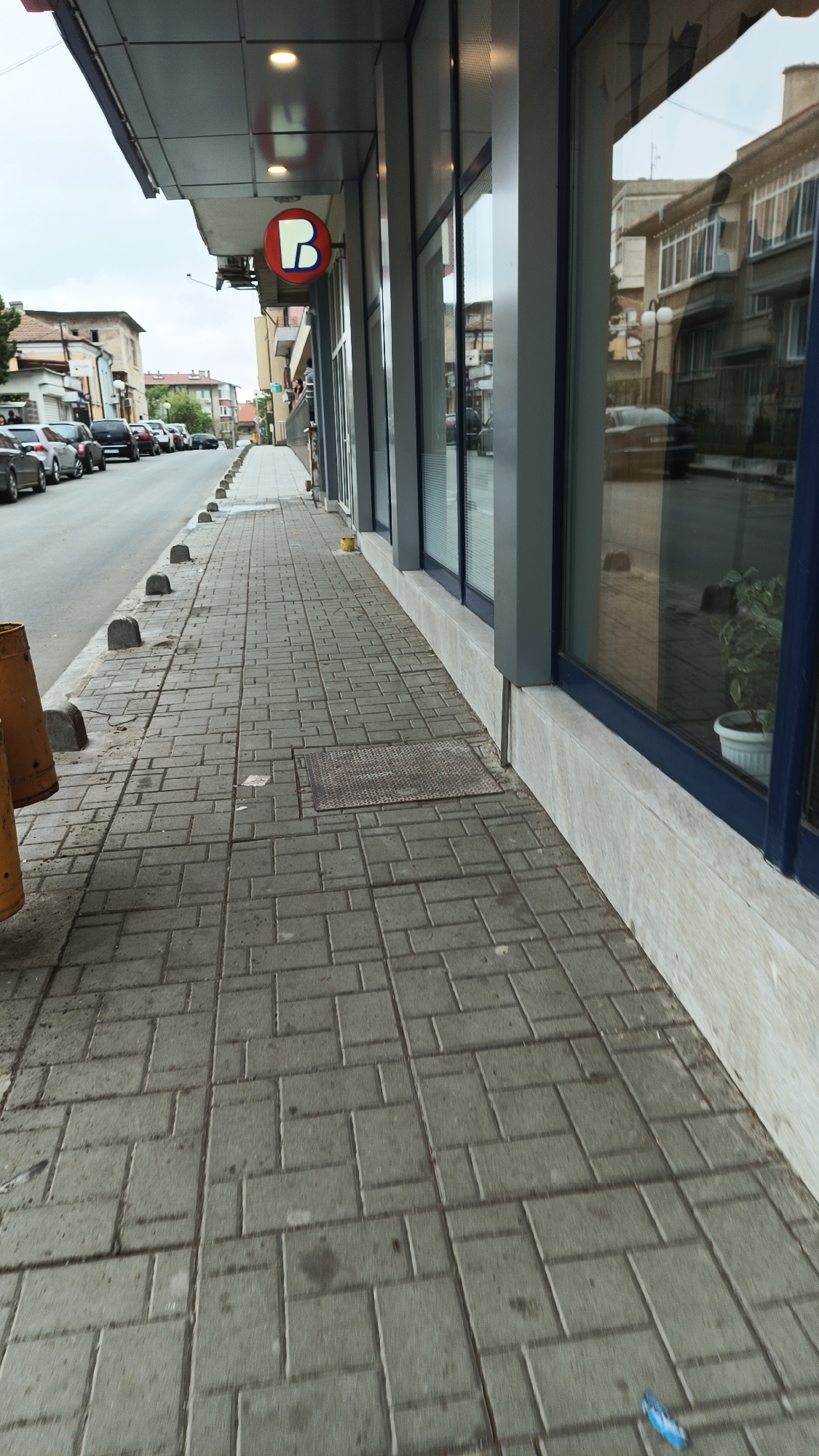 A street scene in Karnobat, Bulgaria, shows a grey-paved sidewalk adjacent to a commercial building with large windows. A circular sign with a white "B" on a red background is visible above the storefront. A row of bollards separates the sidewalk from the street, where several parked cars are visible. The building's facade features grey framing around the windows, which reflect the buildings across the street. A plant in a white pot is visible inside near the window. The sky is overcast. A metal grate is embedded in the sidewalk.