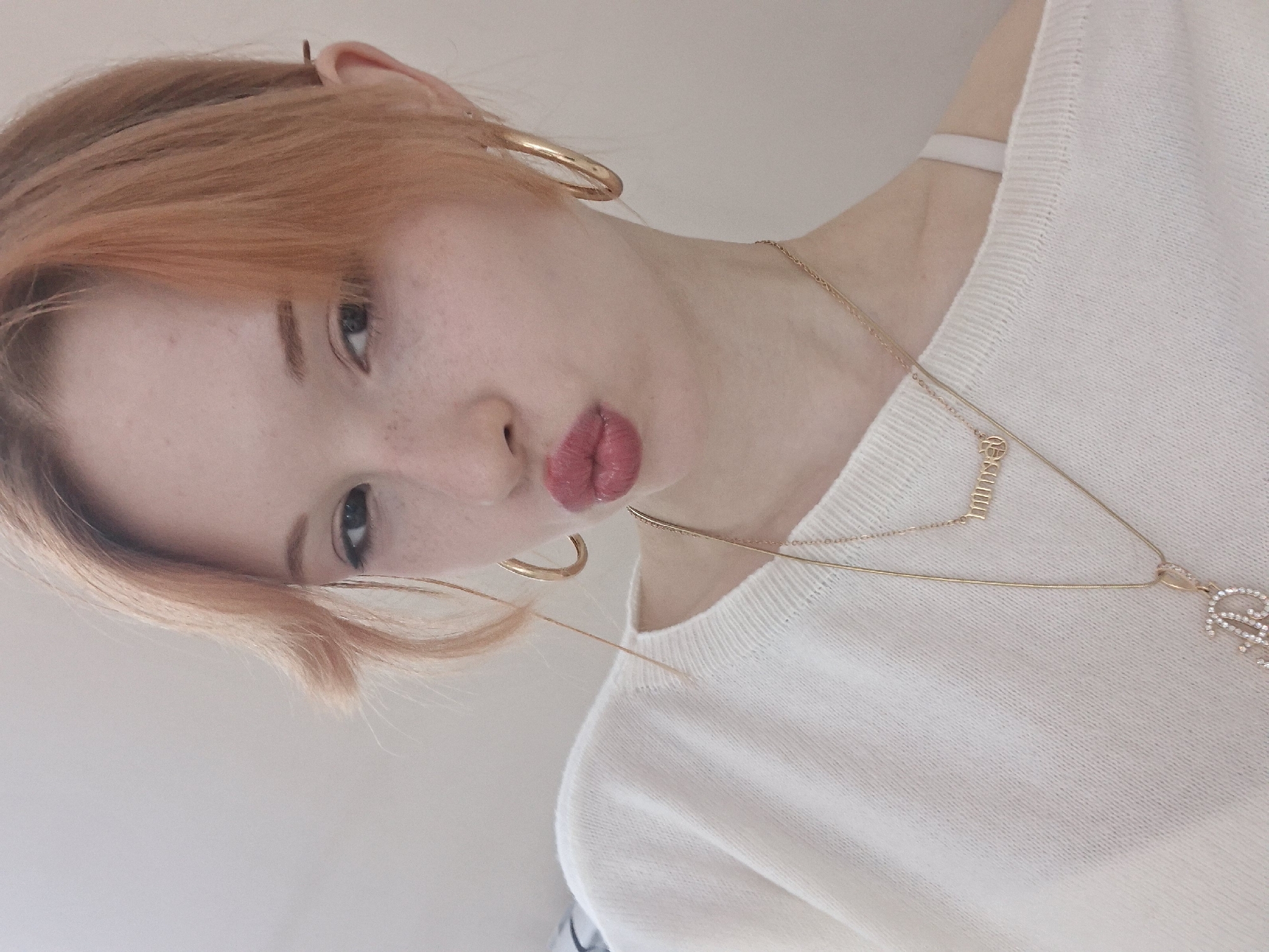 A close-up portrait, taken with a slight upward angle and tilt, features a person with fair skin and light ginger hair. The individual looks directly at the camera with a confident, playful expression, highlighted by slightly pouted and glossy pink lips. Subtle freckles are visible across the bridge of the nose and cheeks. The person wears large, gold-toned hoop earrings and two layered gold-colored necklaces. The shorter necklace bears a small rectangular pendant clearly engraved with the word "BOSS." The longer necklace features a more prominent, ornate pendant adorned with small shimmering details, resembling a stylized initial, possibly 'R' or 'RS'. The subject is dressed in a textured, light-colored, likely white or off-white, knitted sweater. The background is a simple, uniformly light-toned wall or surface, suggesting an indoor setting with soft, diffused lighting. The image captures the upper torso and head of the subject. This scene is situated in Chaikovsky, Russia.