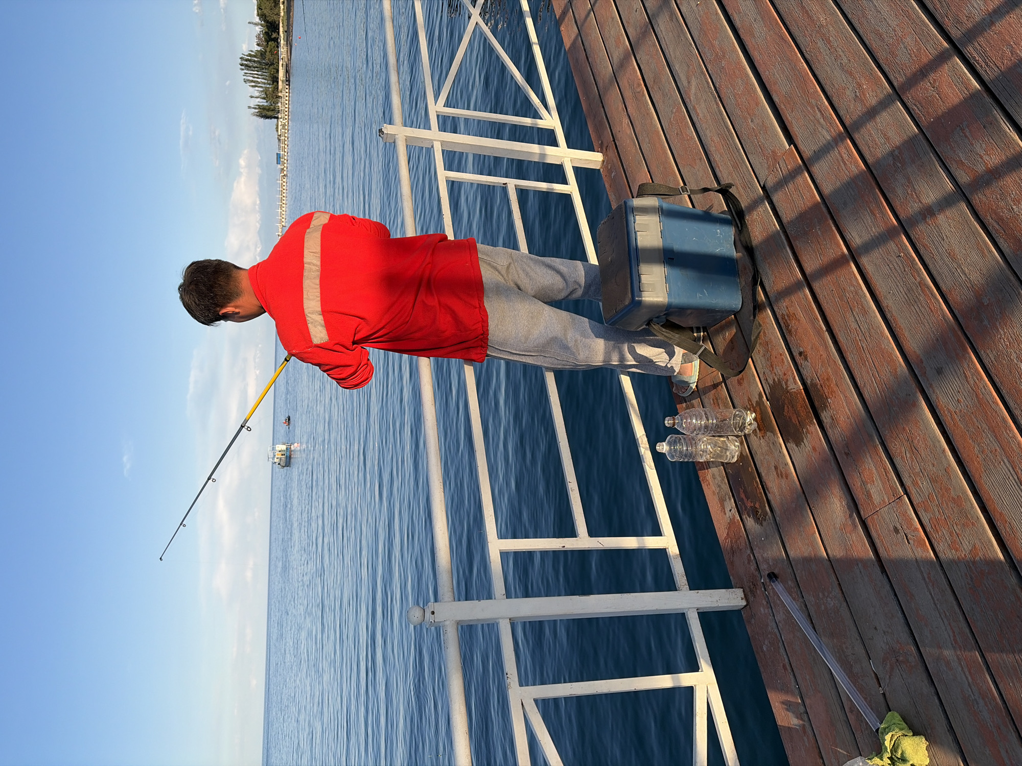 The image depicts a person fishing from a wooden pier. The person, seen from behind, is wearing a red jacket and grey sweatpants and is holding a fishing rod, casting into the calm blue water. Beside them on the pier are two empty plastic water bottles and a blue and grey fishing tackle box. A white railing lines the edge of the pier. In the background, the water extends to the horizon, with a few boats visible in the distance. The sky is a clear blue with a few wispy clouds. The overall scene suggests a peaceful outdoor activity in a waterfront setting.