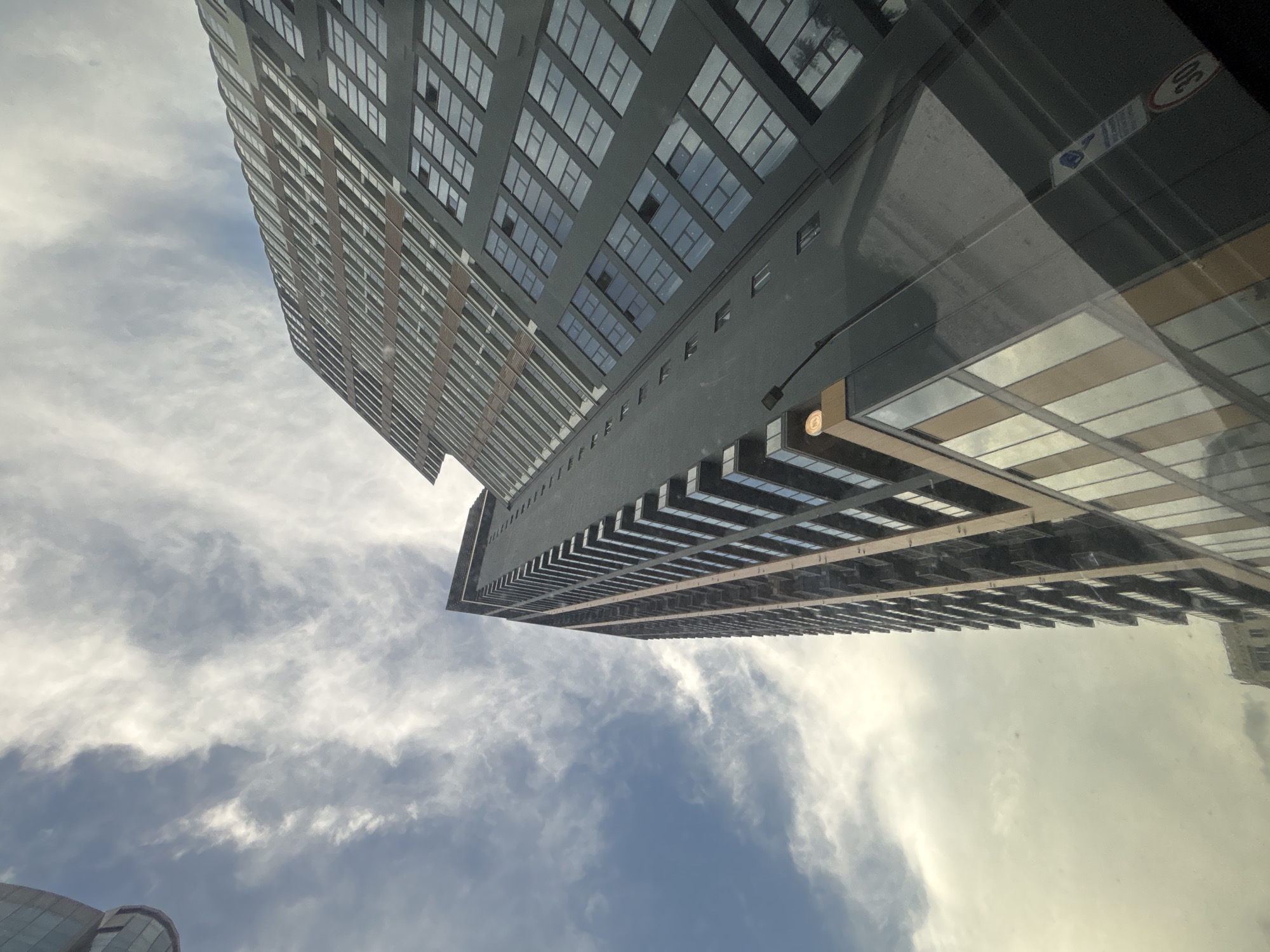 A low-angle shot captures two tall buildings against a backdrop of a cloudy sky. The buildings are predominantly gray with repetitive window patterns. One building is positioned diagonally across the upper left portion of the frame, while the other spans horizontally in the right half. The sky is filled with white and blue clouds. The location is most likely Tbilisi, Georgia, based on the architectural style.