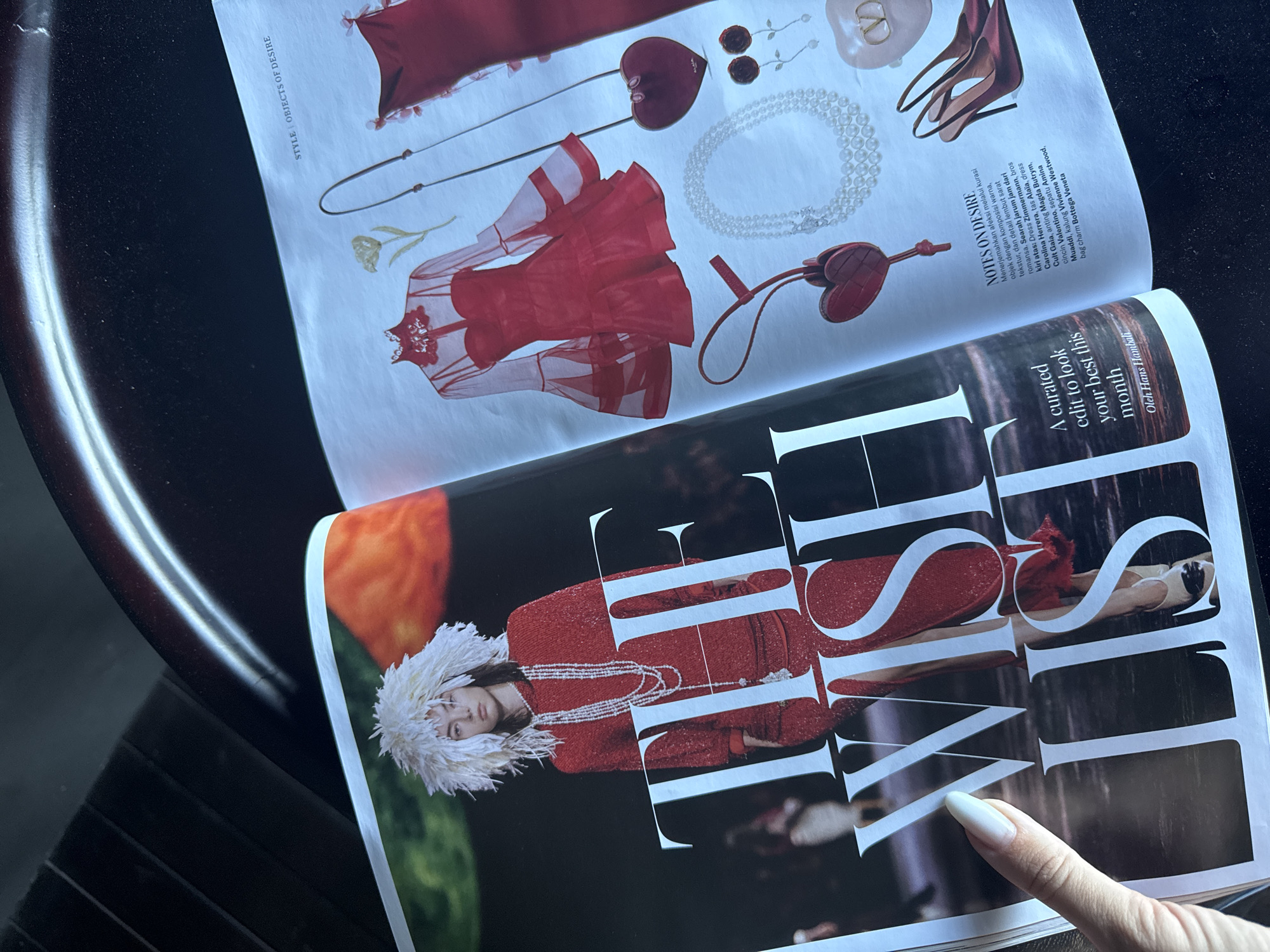 The image displays an open fashion magazine angled on a dark, reflective surface, likely a table. A person's finger, with a long white manicured nail, points towards the bottom right page.
The magazine’s right-hand spread, titled "STYLE | OBJECTS OF DESIRE," showcases a flat lay of red and pearl-themed fashion items. These include a sheer red long-sleeved dress, a solid red dress, a long multi-strand pearl necklace, heart-shaped earrings, a heart-shaped bag charm, red high-heeled shoes, and a subtle golden flower illustration. Adjacent text under "NOTES ON DESIRE" describes these "objects of desire" in Indonesian, listing brands like Zimmermann, Carolina Herrera, Alaïa, Cult Gala, Magda Butrym, Valentino, Amina Muaddi, and Vivienne Westwood.
The left-hand page features a stylish model wearing a red sparkling outfit, multiple pearl necklaces, and a distinctive white feathery hat. Overlaying the image of the model are large, elegant white letters spelling "THE LIST." Below this, the text reads "A curated edit to look your best this month," credited to "Oleh Hans Hambali." The overall mood is sophisticated and fashion-focused, with strong visual emphasis on red, white, and black tones. The lighting is bright and even, illuminating the pages and creating subtle reflections on the glossy dark background.
