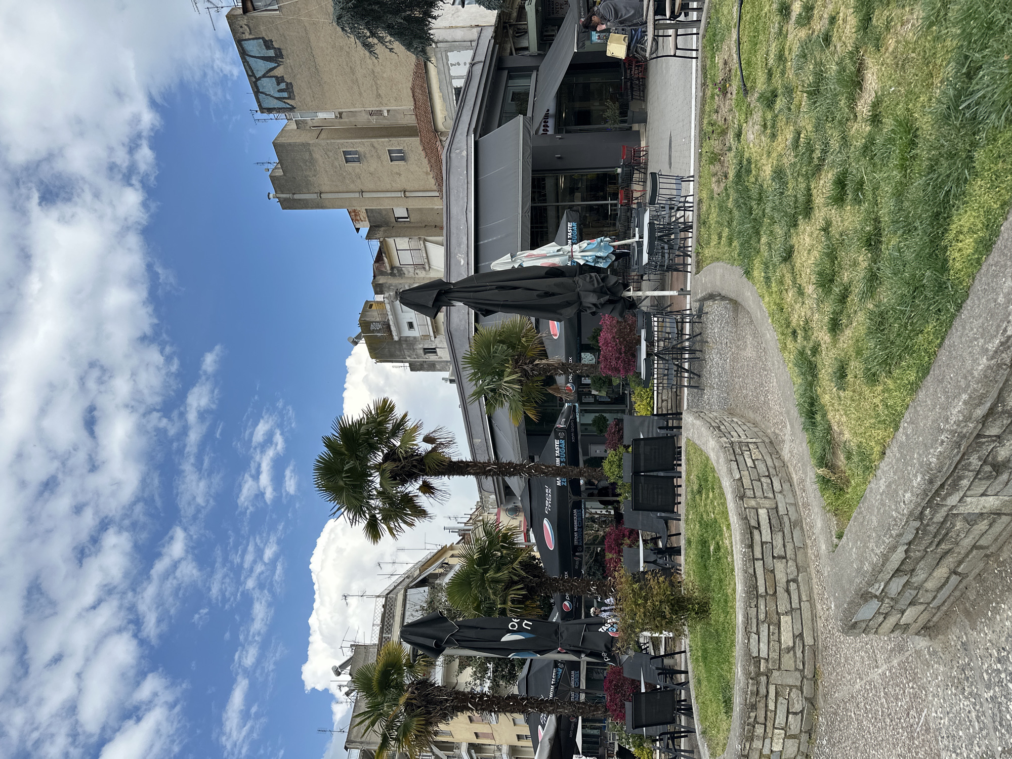 Here is a forensic description of the image:
The image depicts an outdoor scene likely in Serres, Greece, given the Greek text visible on some of the objects. The sky is blue with scattered white clouds. The foreground features a low wall constructed of bricks, and behind it is a grassy area. Further back, an outdoor seating area is set up with tables, chairs, and closed black umbrellas with branding in Greek and the word "TASTE" are apparent. Several palm trees are situated among the seating. The buildings are multi-story and of varied architectural styles. A man is seen sitting at a table near a store named "Tappino".