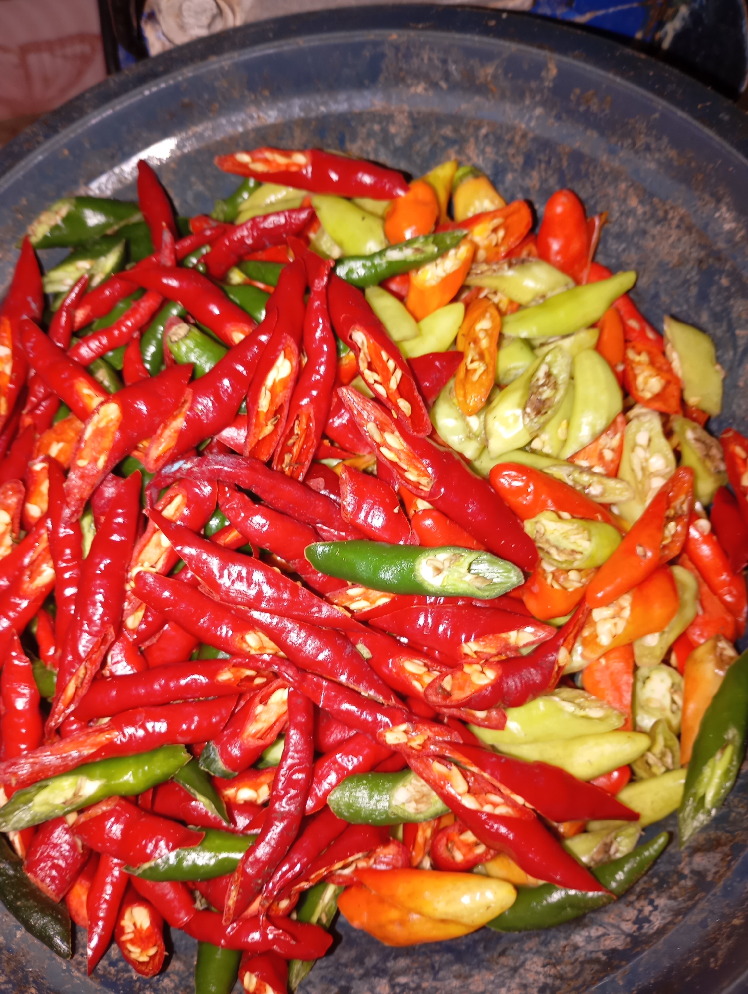 The image shows a close-up view of a dark blue plastic basin filled with sliced chili peppers. The chilies are a mix of red, green, and orange, and they appear to be freshly cut, with visible seeds and inner flesh. Some chilies are whole and some are halved or cut into smaller pieces. The basin is weathered and appears to be located outdoors, possibly in a rural setting. The background is blurred but suggests a natural environment. The lighting is dim, and the image quality is grainy, indicating it was likely taken with a low-resolution camera or in low light conditions. It is not possible to determine the specific location within Kalitinggar, Indonesia from the image alone.