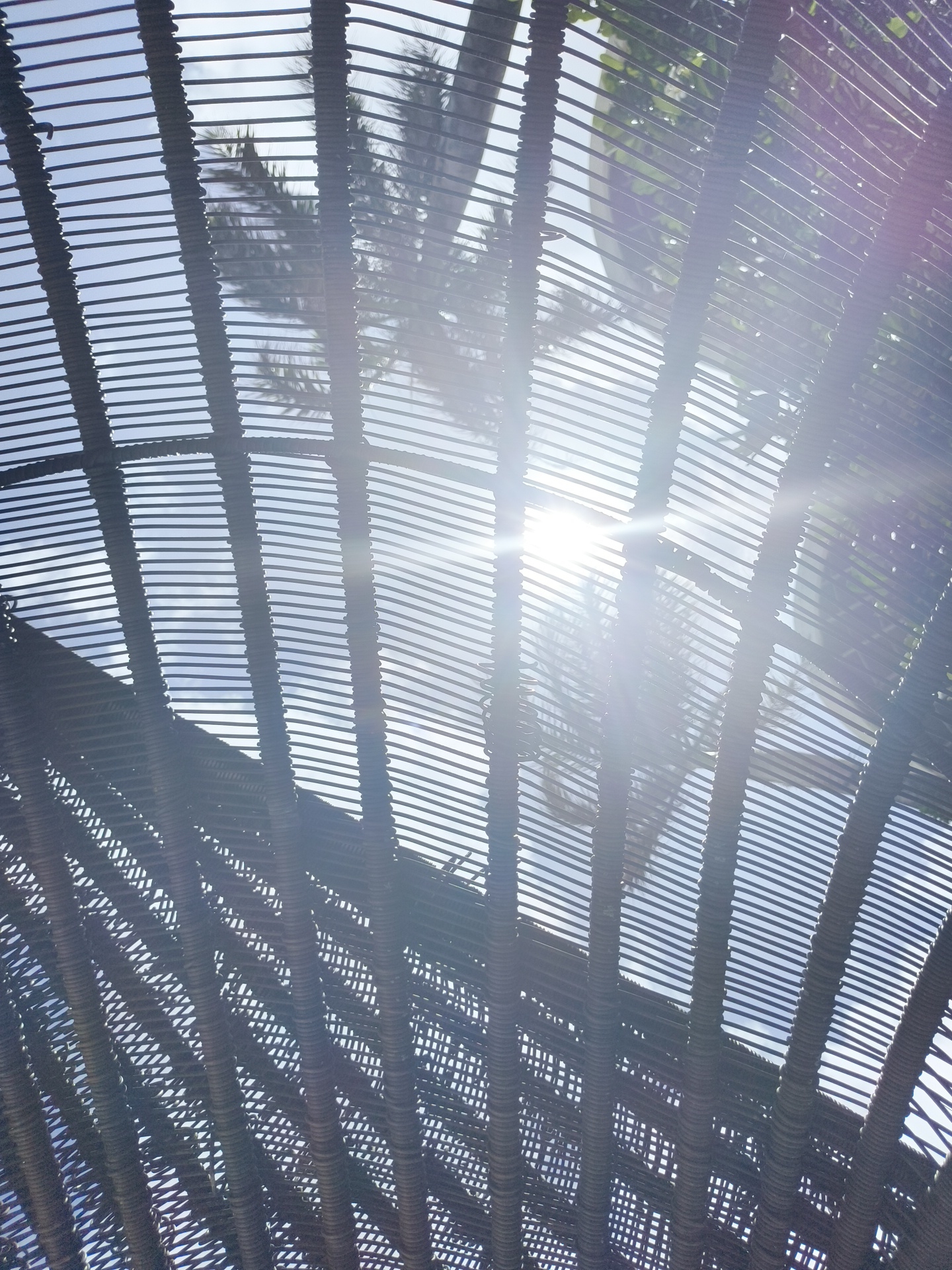 The image depicts a view looking upwards through a woven, open-air structure, possibly a roof or shade element, likely made of natural materials like rattan or bamboo. The structure is composed of numerous thin, horizontal strands woven around vertical supports, creating a striped effect. Through the gaps in this weaving, a bright sun is visible, creating a strong lens flare and casting light patterns. The sky is a pale blue, and the blurry silhouettes of palm fronds can be seen against it, suggesting a tropical or sub-tropical environment. This scene could be found in an outdoor seating area, a garden pavilion, or a beachside establishment in Phú Quốc, Vietnam, a location known for its tropical climate and natural beauty. There are no people or explicit actions depicted. The focus is on the interplay of light, shadow, and the textured materials of the structure against the sky and foliage.