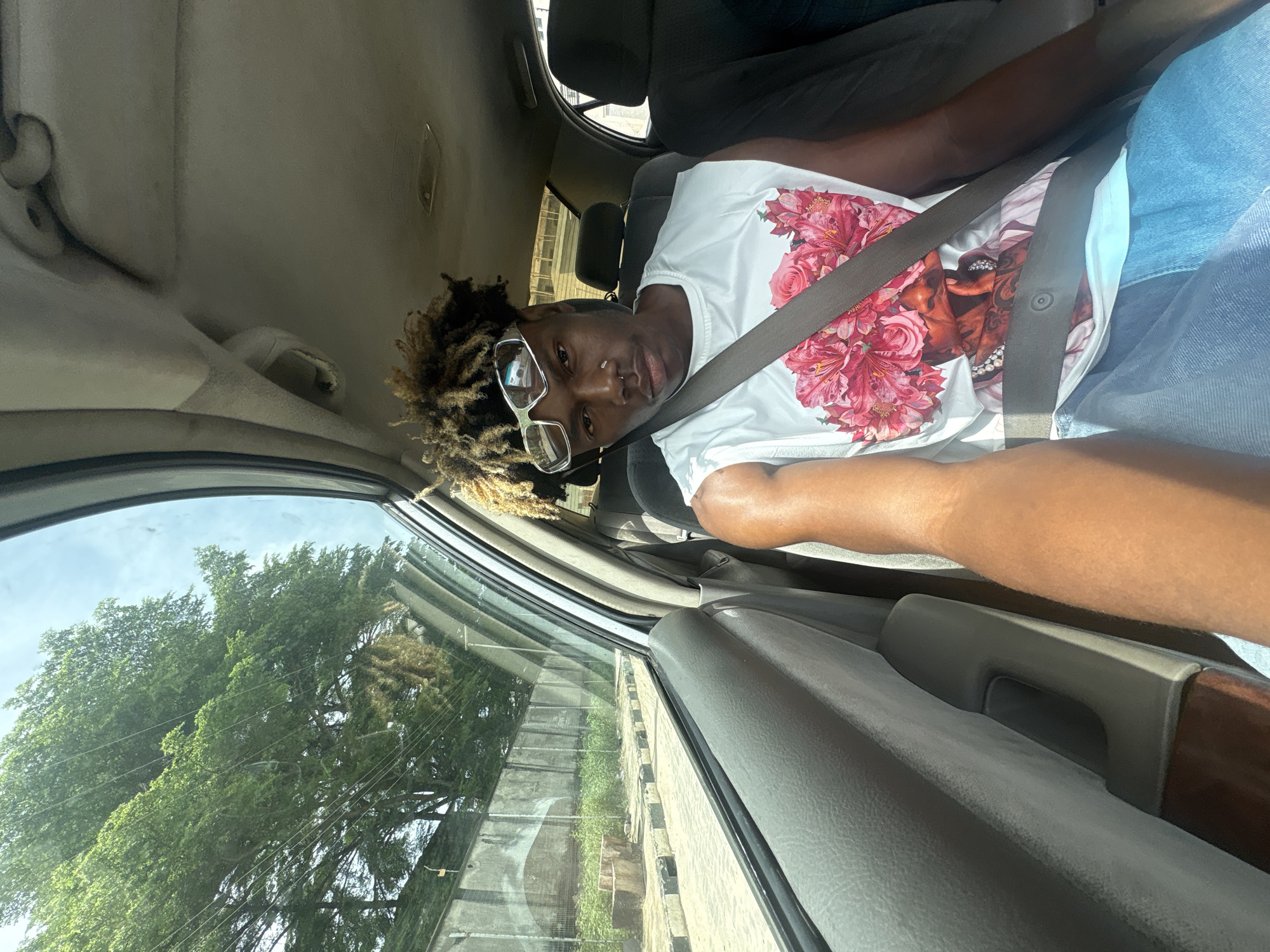 A person with dreadlocks and glasses is sitting in the passenger seat of a car, wearing a seatbelt and a white t-shirt with a floral and face print. The car is moving, as evidenced by the trees and a wall passing by outside the window. The image suggests a daytime setting. The person is looking at the camera. Igboefon, Nigeria, is indicated as the location.