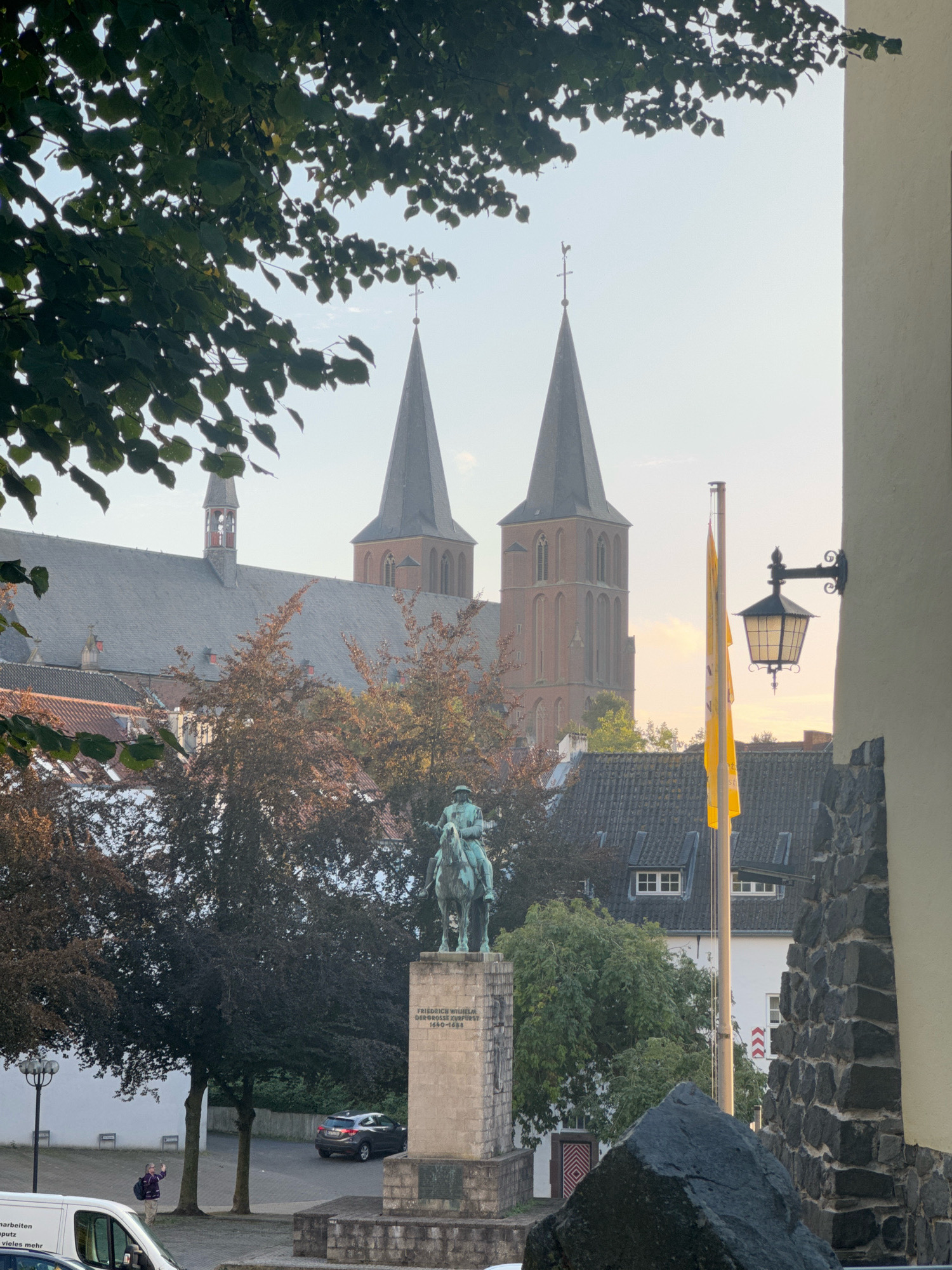 The image shows a view of the St. Maria Magdalena church in Cleves, Germany, as seen from a street. The church has two prominent spires. In the foreground, there is a statue of a man on horseback on a pedestal. The pedestal has an inscription that reads "FRIEDRICH WILHELM DER GROSSE KURFURST 1620-1688." There is a yellow flag on a flagpole to the right of the statue. Trees with reddish-brown leaves are visible between the statue and the church. A street lamp and a parked car are also in the scene. The sky is clear and overcast, with the sun appearing to be low in the sky, suggesting it is either early morning or late afternoon. A person is waving from behind a white van in the lower left corner of the image.