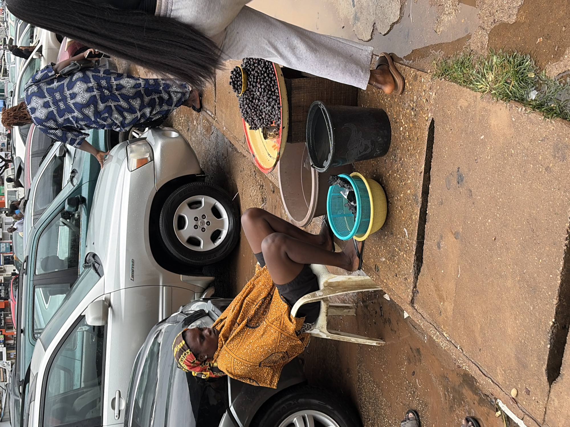 The image depicts a street scene in Jos, Nigeria, on a rainy day. In the foreground, a woman sits on a white plastic chair, draped in an orange and yellow patterned fabric, and wearing black shorts and flip-flops. She is looking down and to her right. In front of her, on a raised wooden platform, are several containers of dark berries, including a large yellow bowl with a green sieve on top, a black bucket, and a large shallow tray overflowing with berries. A brown basin filled with water is also visible.
Behind the woman, a silver SUV is parked, partially obscuring a dark-colored car. Further in the background, more vehicles are parked, and the architecture suggests an urban environment. Another woman, wearing a blue and white patterned dress, is seen walking away from the viewer towards the vehicles. A person wearing light grey sweatpants and sandals is standing near the berries, with their back to the camera. The ground is wet and muddy, indicating recent rainfall. The overall impression is of a marketplace or roadside stall operating under inclement weather.