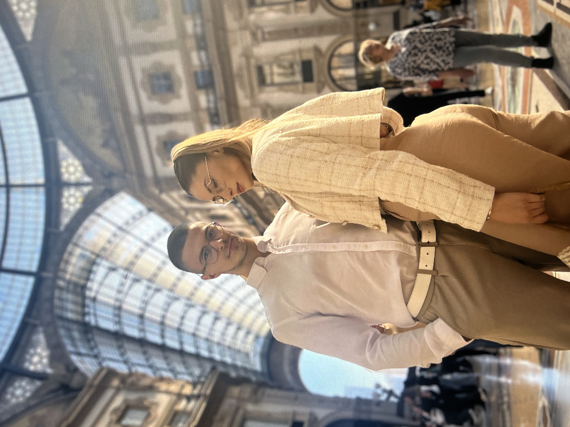 A sideways, medium shot shows a man and a woman posing together in Galleria Vittorio Emanuele II in Milan, Italy. The man is in the lower part of the shot. He has short, dark hair and wears round, dark glasses, a white button-down shirt, khaki pants, and a white belt. The woman, posed behind him, has long blonde hair and wears metal framed glasses, a cream-colored jacket, and khaki pants. Behind them, other people are milling around. The glass domed roof of the galleria is visible.