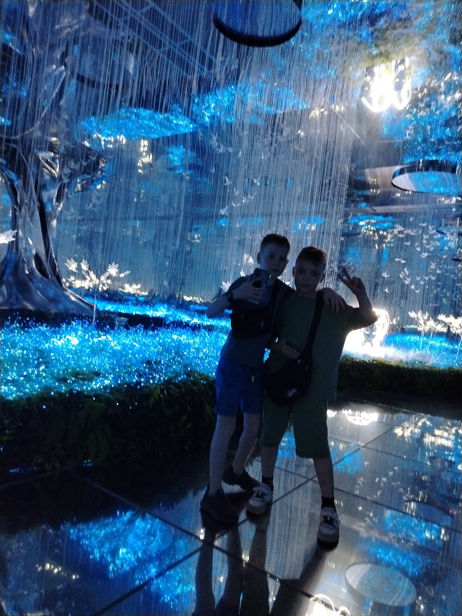 Two young boys are posing for a picture inside what appears to be an indoor display decorated in glowing blue lights. The boy on the left is holding a phone and has his arm around the other boy. The boy on the right is holding up a peace sign. The surrounding environment includes a large artificial tree, glowing flora on the ground, and string lights hanging from the ceiling. The ground is reflective and looks like a polished floor. Based on the location inferred, this is likely at the Kazan Family Center, a unique building complex in the city of Kazan, Russia.