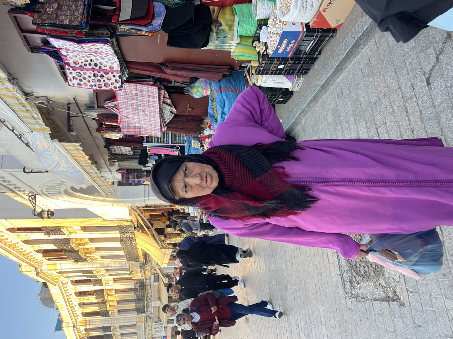 The image captures a bustling street scene in the city of Tanger, Morocco, likely within a market district. In the immediate foreground, an elderly woman, appearing to be in her 60s or 70s, stands centrally, looking directly forward with a neutral expression. She wears a vibrant deep purple djellaba with vertical embroidery, a black headscarf, and a red and black plaid fringed scarf wrapped over her head. Her right hand is placed on her hip, and her left hand holds a blue plastic shopping bag and another light-colored bag.
To the woman's right, a market stall is visible, displaying various merchandise including numerous patterned reusable shopping bags hanging from a dark brown panel, black skullcaps, and bulk items such as white sacks of grains or legumes, crates of garlic, and a large blue sack containing colorful contents, arranged on the paved ground. Above this stall, a white building segment features an arched entrance and a yellow-and-white striped awning, with a decorative black lantern affixed to the wall.
To the woman's left and extending into the background, a light-colored paved pedestrian street is populated with multiple individuals. A man in a red sweater and dark trousers walks towards the left mid-ground. Other pedestrians are visible further back. The backdrop on the left features a prominent, multi-story building characterized by a cream-yellow facade, arched windows, balconies with dark railings, and ornate architectural detailing, illuminated by bright sunlight. The sky above is clear and blue.
