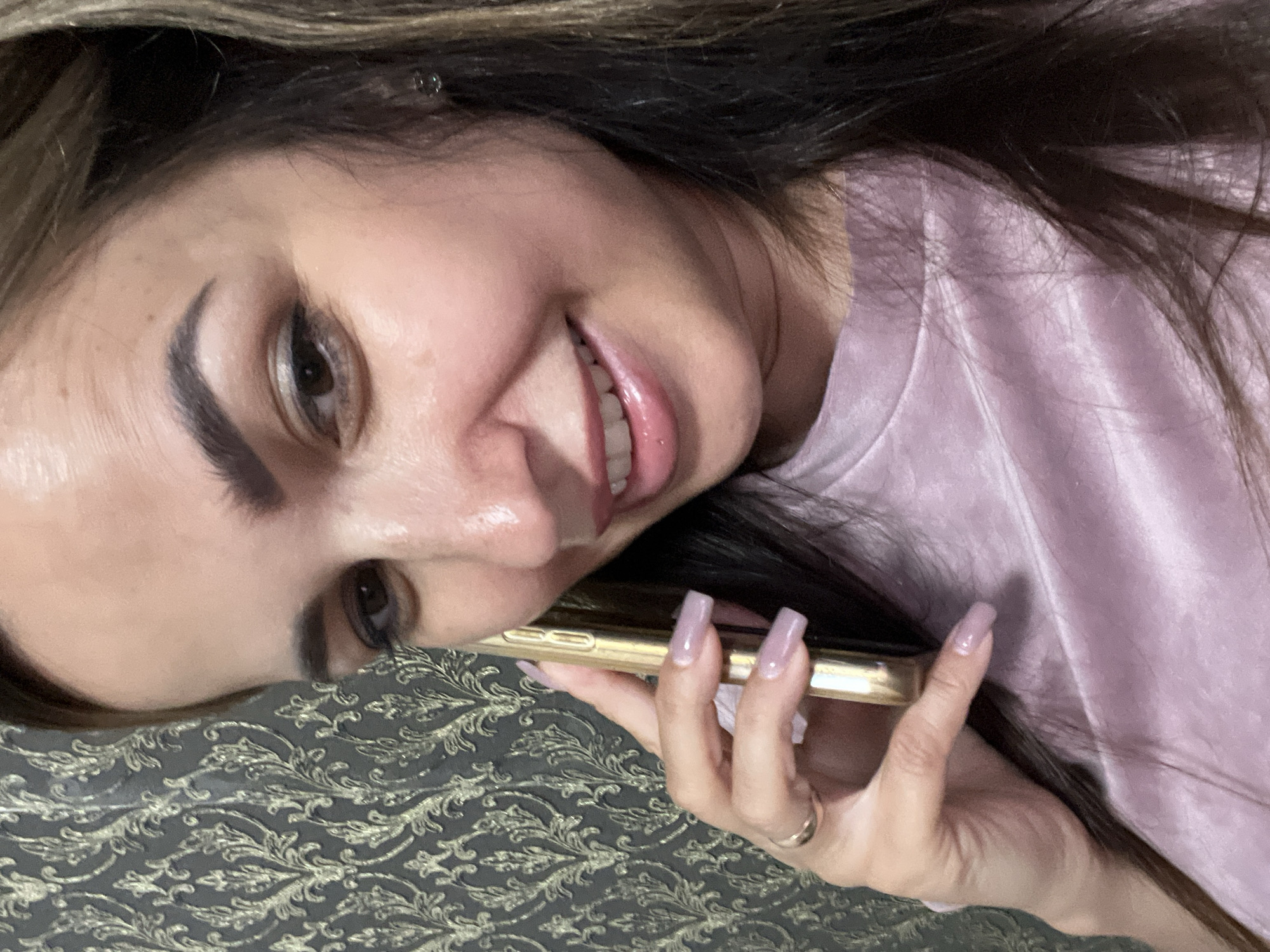 A woman is shown from the chest up, holding a gold smartphone to her ear. She is looking towards the camera with a smile and appears to be in a casual setting, possibly at home. The background features a textured wallpaper with a damask pattern. She is wearing a pink, silky top and has long, dark hair with highlights. Her nails are painted a light purple. A gold ring is visible on her left hand. The lighting suggests an indoor environment.