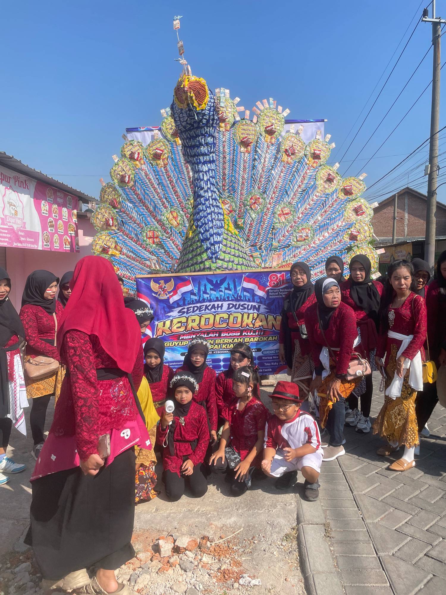 In Kebon Waris, Indonesia, a vibrant scene unfolds under a clear blue sky, likely during a daytime celebration. The focal point is an enormous, elaborately constructed peacock float, its tail feathers meticulously crafted from numerous small, colorful packets, possibly snacks or candies. The peacock stands tall, dominating the background, with its head and neck adorned with similar colorful packaging, creating a striking visual.
In front of the float, a diverse group of people are gathered, many dressed in traditional red and black attire. Women in elegant red tops and patterned skirts, some with headscarves, are positioned in the foreground and middle ground. Several children are also present, some in matching red outfits and headwear, adding to the festive atmosphere. One young girl holds a small white device, possibly a fan or a toy. A young boy in a red and white shirt and shorts, wearing glasses and a red hat, is crouched down, seemingly observing the scene.
The environment appears to be a street or an open area within the city. To the left, a building with pink signage, possibly a shop or establishment named "Dapur Pink," is visible, with further signage displaying food items. The ground is paved with cobblestones and has some scattered debris. Utility poles with wires are present in the background, indicating an urban setting. A banner behind the peacock float bears text in Indonesian, featuring Indonesian flags and the number "81," suggesting a significant event or anniversary. The overall impression is one of a community event, possibly a parade or festival, characterized by its colorful decorations and the participation of its residents.