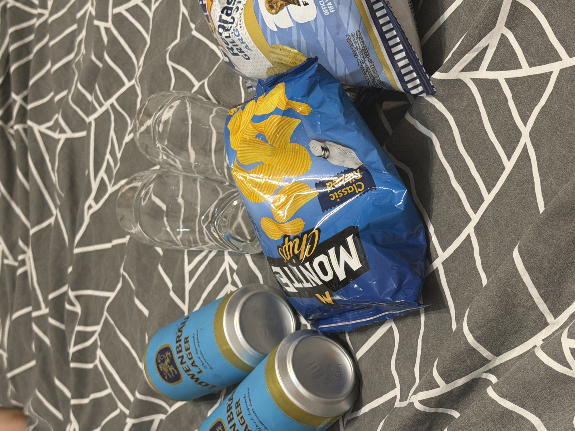 Two cans of Lowenbrau Lager are placed on a grey and white patterned fabric, one partially obscured by the other. To the right of the cans, two empty glass cups are positioned. In the foreground, two bags of potato chips are visible: a blue bag labeled "MONTE Chips Classic" and a lighter blue bag with a FIFA World Cup graphic. The scene appears to be indoors, possibly on a bed, in Budva, Montenegro. No people are present in the image.