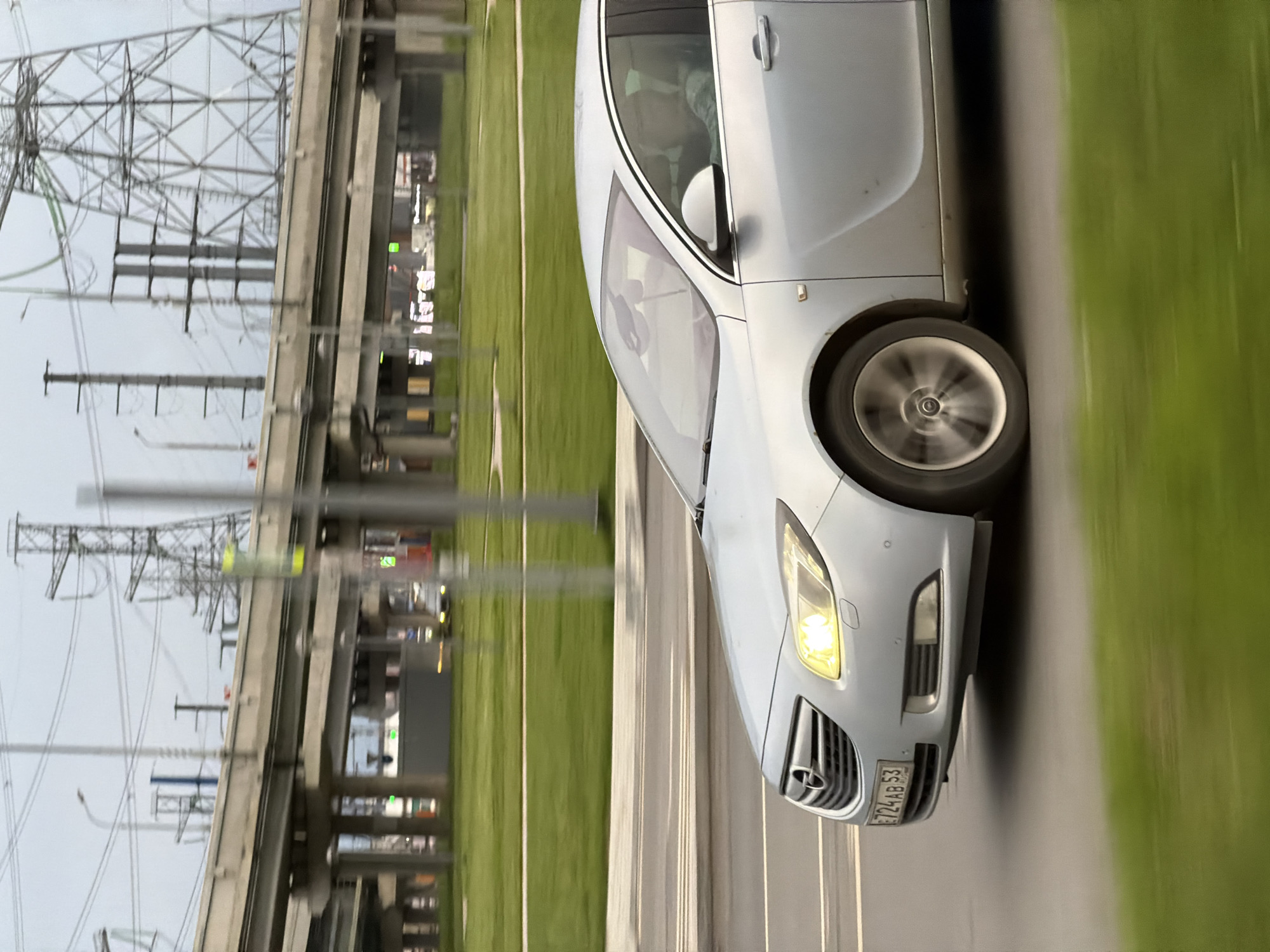 The image shows a silver car driving on a road in an urban area. The license plate of the car is visible, displaying "E724 AB 53". The background shows a grassy area and a complex infrastructure of overpasses and power lines. The location is in the city of Unknown, Unknown.