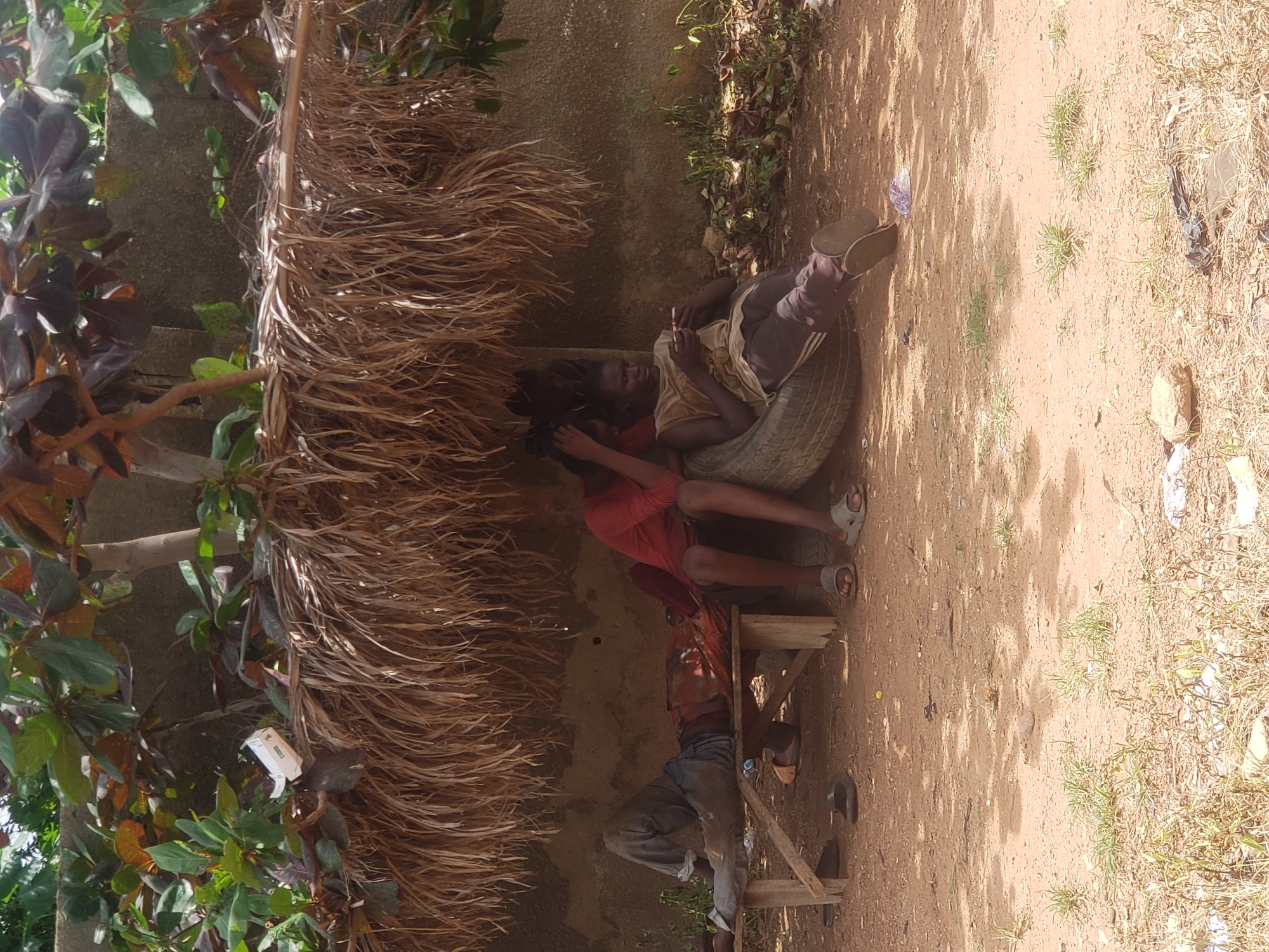 In Narayi, Nigeria, three individuals are positioned under a thatched roof structure. One person, dressed in a red long-sleeved shirt and dark shorts, is seated. Another person, wearing a sleeveless tan shirt and dark pants, is sitting on a tire and appears to be looking at a phone. A third individual, clad in a red patterned shirt and gray pants, is lying down on a wooden bench. The ground is covered in sand and dry grass. The environment includes some trees with green and reddish-brown leaves above the thatched roof, and a concrete wall behind the structure. Scattered debris, including plastic bags and wrappers, is visible on the ground.