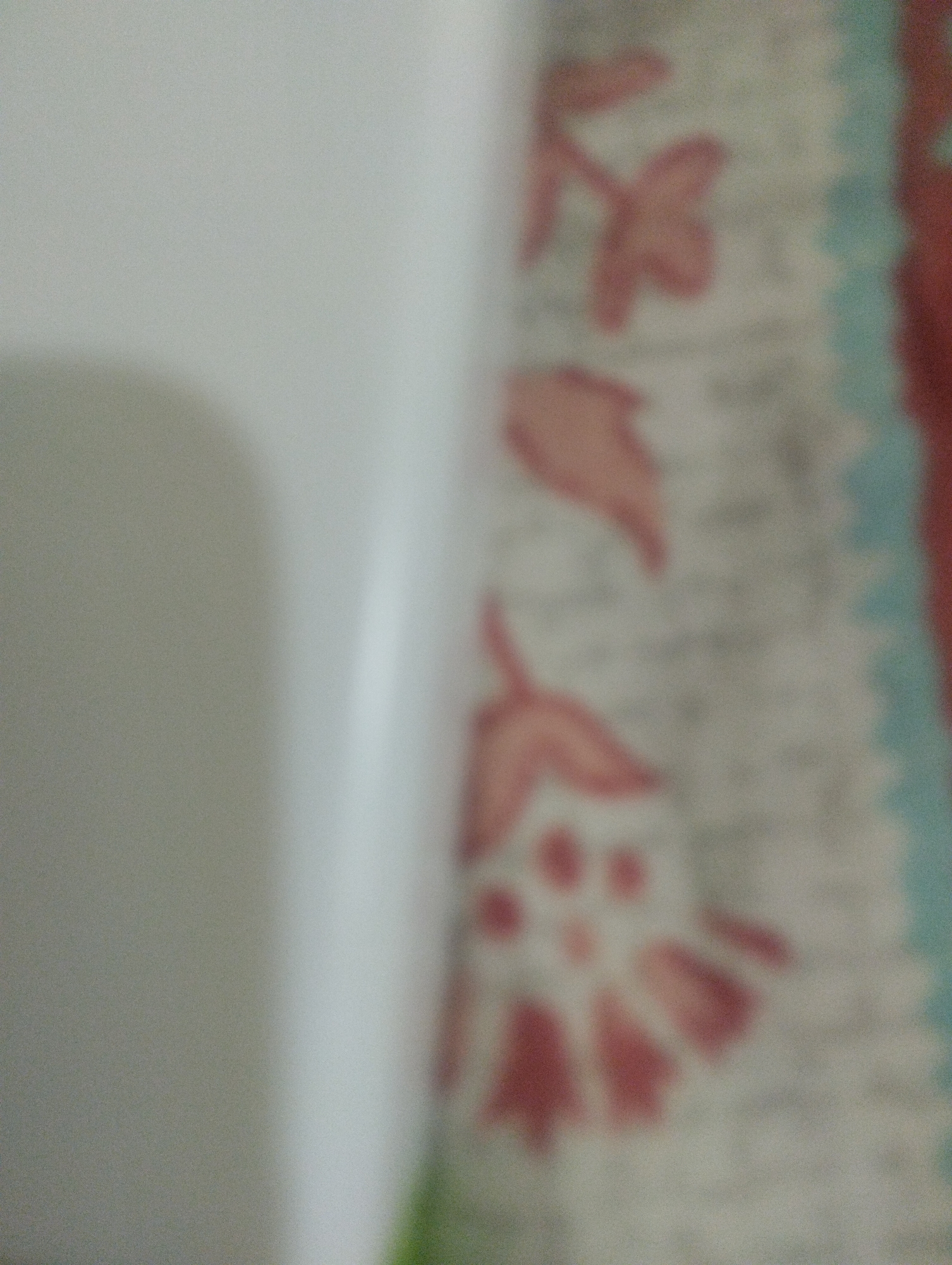 The image shows a close-up view of a patterned fabric with a lighter object on the left side. The fabric has a repeating pattern of stylized pink flowers on a light background, bordered by a band of light blue and a darker red edge. The lighter object is out of focus and appears to be a smooth, curved surface, possibly a piece of furniture or a container. The overall impression is of a textured surface with a floral design. The image is taken in a slightly blurry fashion.