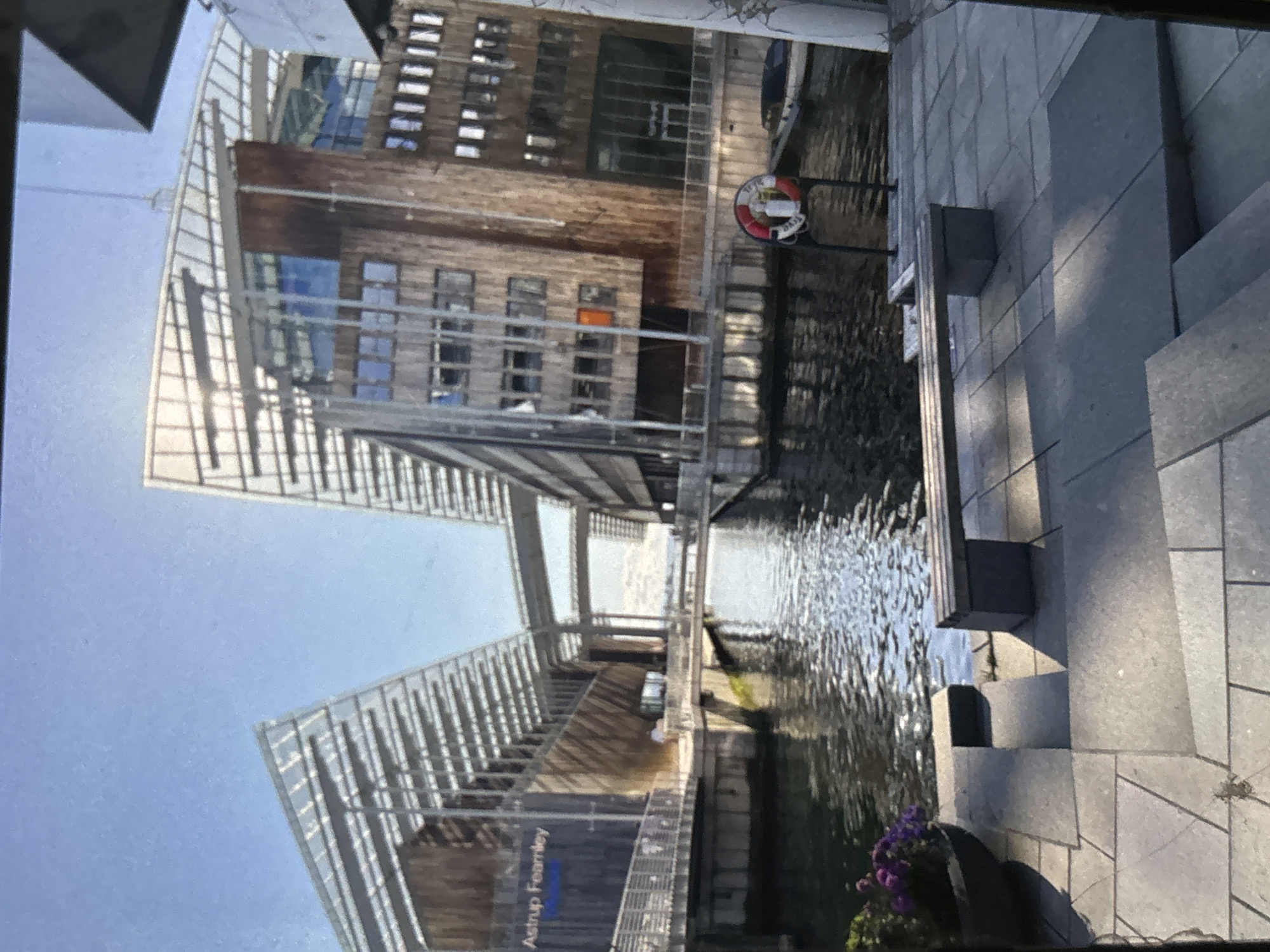 The image depicts a waterfront view of the Astrup Fearnley Museum, a contemporary art museum located in Oslo, Norway. The building features a distinctive modern architectural design with angled glass roofs and wooden facades. It is situated on the edge of a body of water, with a canal or harbor running alongside it. In the foreground, a paved area with stone steps leads down to the water. A wooden bench is placed on this paved area, overlooking the water and the buildings. A potted plant with purple flowers is visible to the left of the steps. In the water, a life preserver is attached to a pole, and a small boat is docked nearby. The sky above is clear and blue. The text "Astrup Fearnley Museet" is visible on the left side of the building.