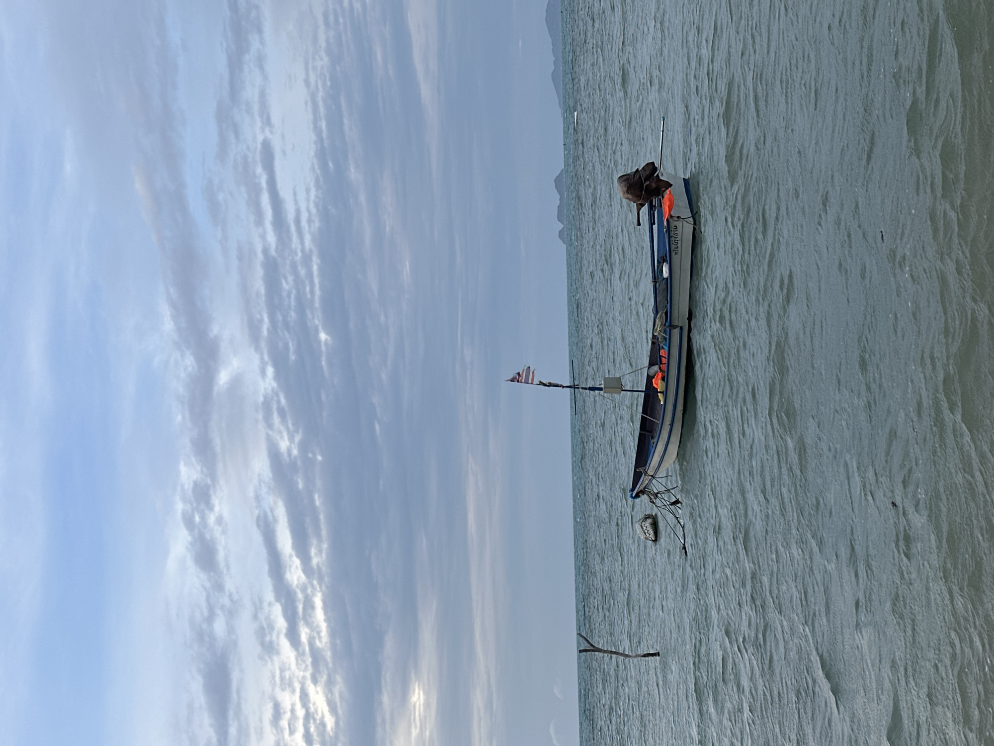 A fishing boat is anchored in choppy, grey water under a cloudy sky. The boat is white with blue trim and has an outboard motor covered with a dark tarp. It flies a small flag and has several orange life vests visible. A wooden post sticks out of the water to the left of the boat, and a buoy is tethered nearby. In the distance, hills are visible on the horizon. The location is the coast of Ko Samui District, Thailand.