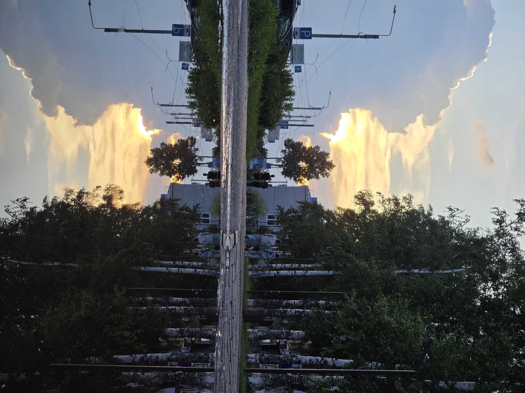 This photograph captures a scene on an overcast day in Chaikovsky, Russia. The image is dominated by a large puddle in the foreground, which reflects the sky, trees, and buildings. To the left of the puddle, a street is visible with a lamppost, traffic signs, and a parked car. Further down the street, two individuals are walking away from the camera. On the right side of the image, a row of birch trees lines the sidewalk, with a building visible behind them. The sky is partly cloudy, with patches of sunlight breaking through the clouds, casting a warm glow. The reflection in the puddle creates a symmetrical and somewhat surreal effect, mirroring the elements of the scene above.