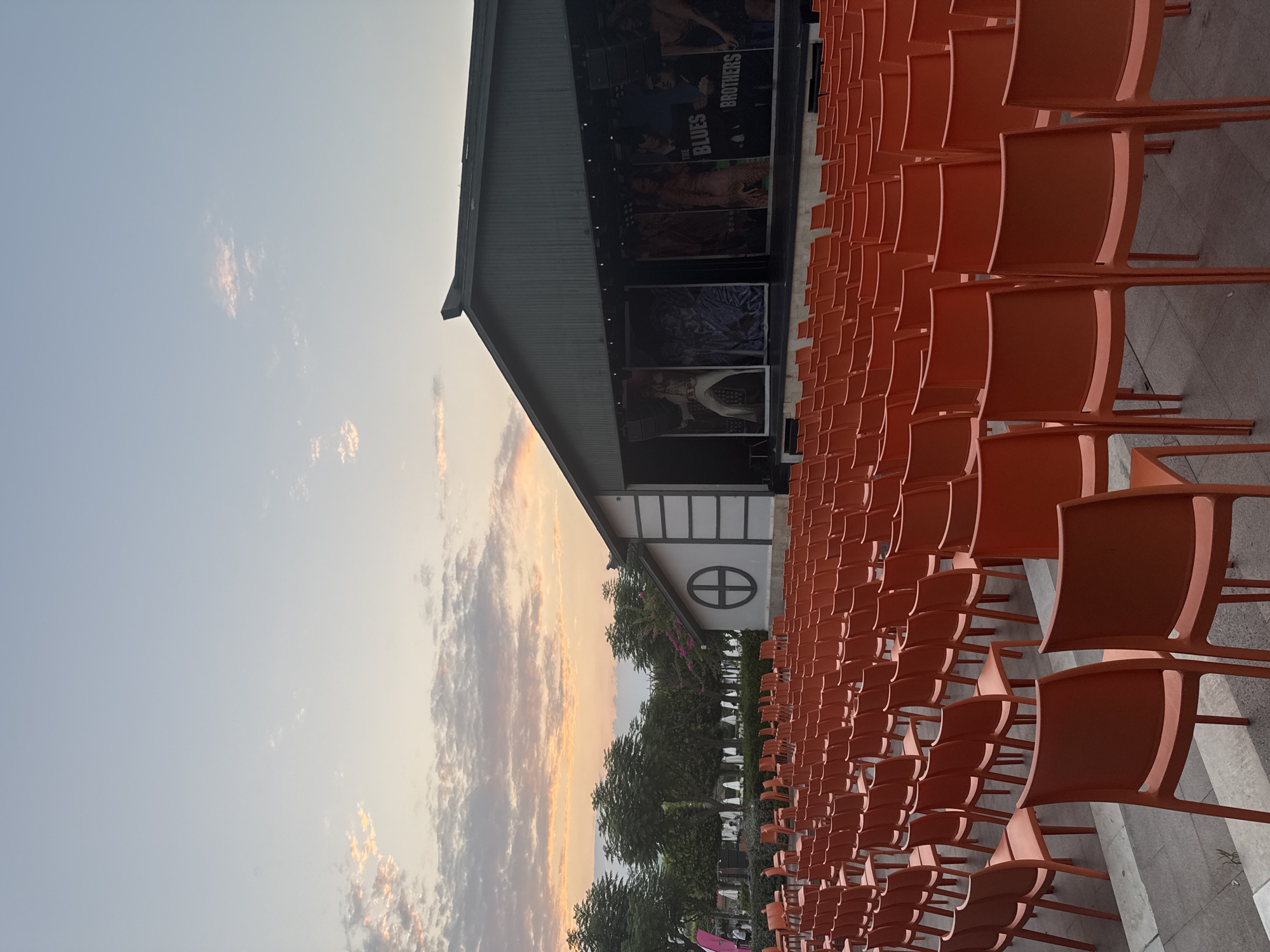 An outdoor amphitheater at dusk in Aksu, Turkey, is depicted. Rows of empty, bright orange plastic chairs face a stage. Behind the stage, banners display images of performers, including one that appears to be Elvis Presley, and another for "The Blues Brothers." The sky above is a pale blue with streaks of soft orange and pink from the setting sun, and scattered clouds. Trees are visible behind the stage area, and a building with traditional Japanese architectural elements, including a circular window, is situated to the left of the stage. The ground in front of the chairs is paved with stone tiles. A few people are visible in the distance near the trees, but the focus is on the empty seating and the stage.