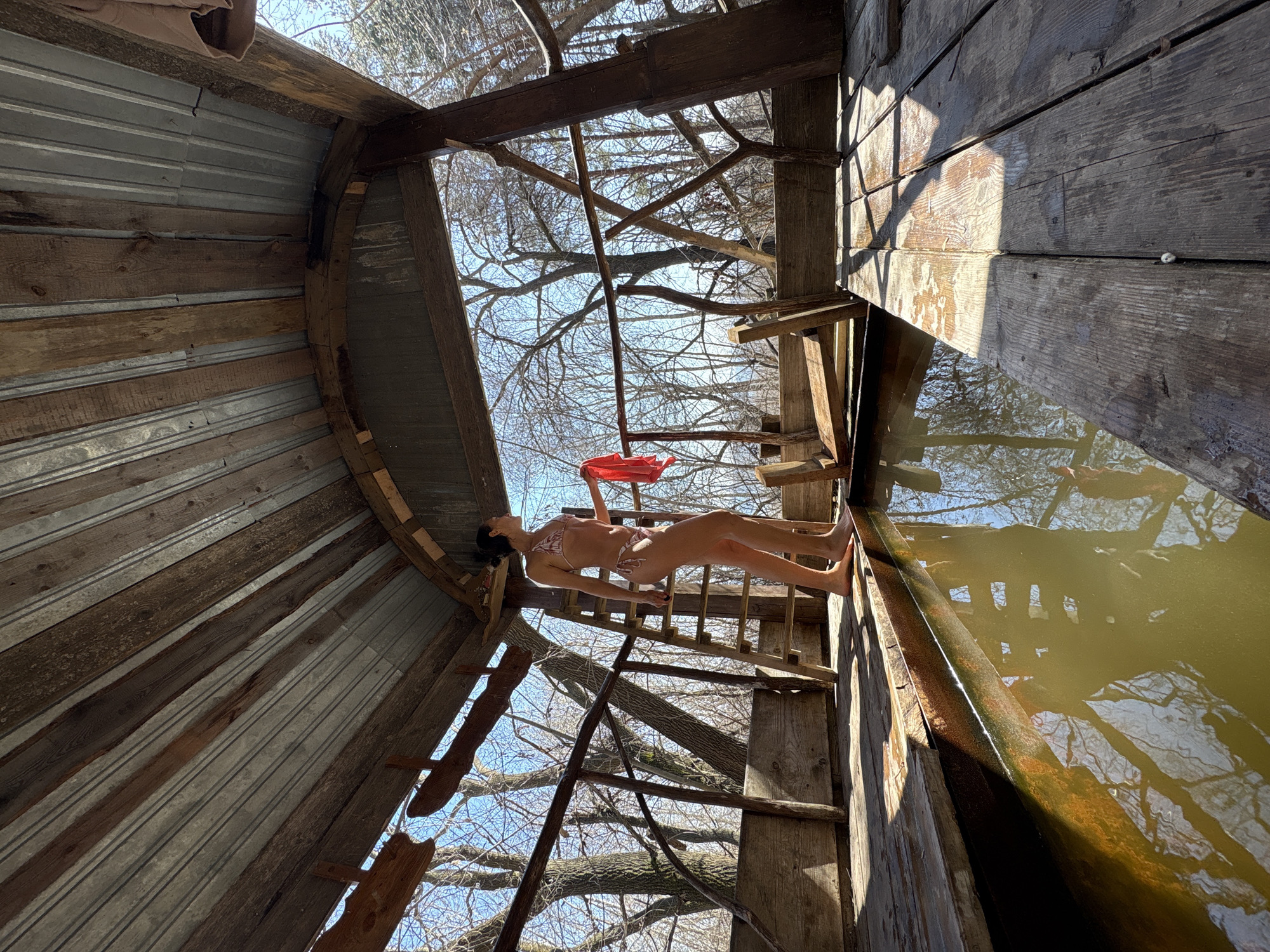 A woman in a bikini lies on a makeshift wooden platform inside a water tower in Ulyanovsk, Russia. The tower’s interior is constructed of wood and corrugated metal, and water fills part of it. The woman holds a red object and appears to be positioned for a photograph. Bare tree branches are visible through the tower's open top.