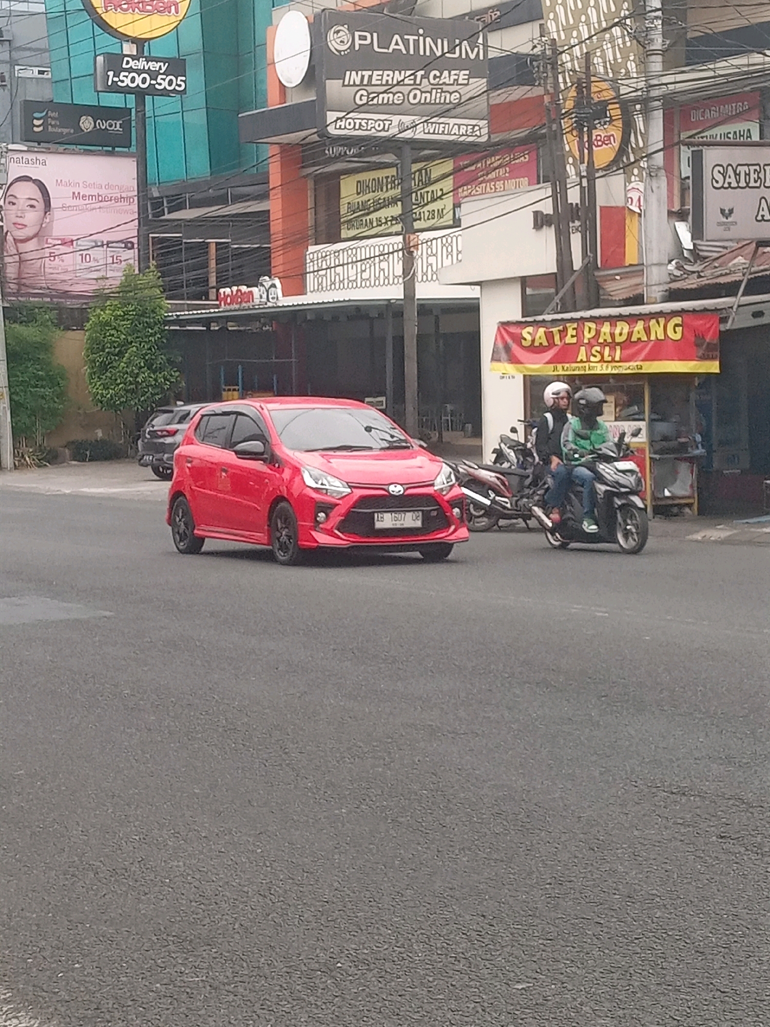 The image shows a street scene in Depok, Indonesia. A red car is driving on the road, with a motorcycle carrying two people behind it. In the background, there are several buildings with various signs, including one advertising "Sate Padang Asli" and another for "Platinum Internet Cafe". There are also trees and power lines visible in the scene. The overall impression is of a busy urban environment.