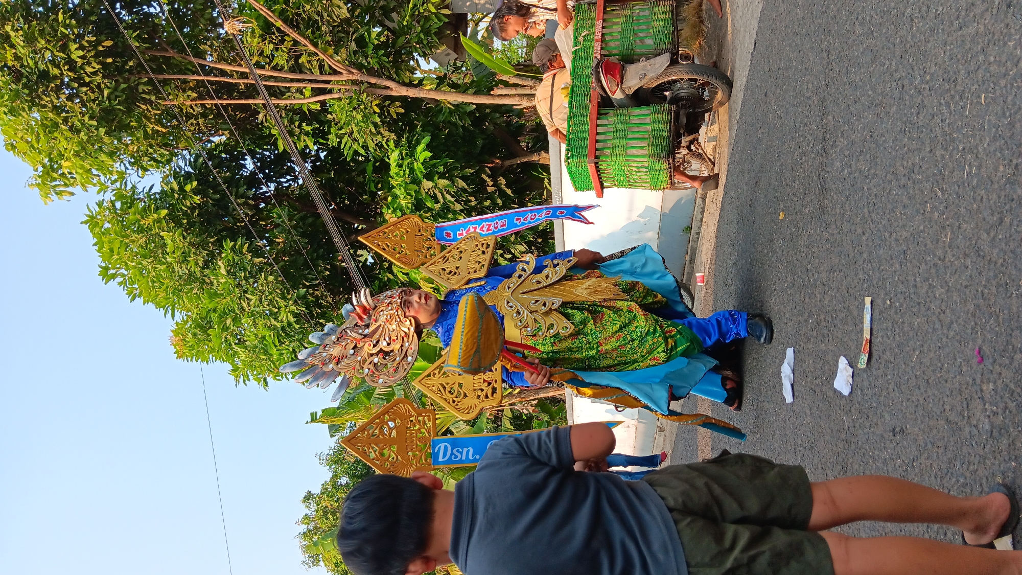 The image captures a daytime scene on a paved street in Cerme, Indonesia, likely during a local festival or parade. The central figure is a person adorned in an elaborate, colorful costume, featuring a large, ornate headdress with feathered accents and golden wing-like decorations extending from the shoulders. The costume includes a blue tunic, green and gold patterned lower garment, and a flowing blue cape. The person holds a large, golden ceremonial object resembling a drumstick or scepter.
To the left of the main subject, a young boy in a grey t-shirt and olive-green shorts stands with his back to the camera, observing the procession. Further in the background, on the right side of the street, a green basket-like container filled with goods is attached to the back of a motorcycle, and two individuals are partially visible tending to it. Behind them, lush green trees and foliage line the street, with utility wires visible overhead. The ground is asphalt, with several discarded white papers and a colorful object lying on it. The overall atmosphere appears festive and communal.