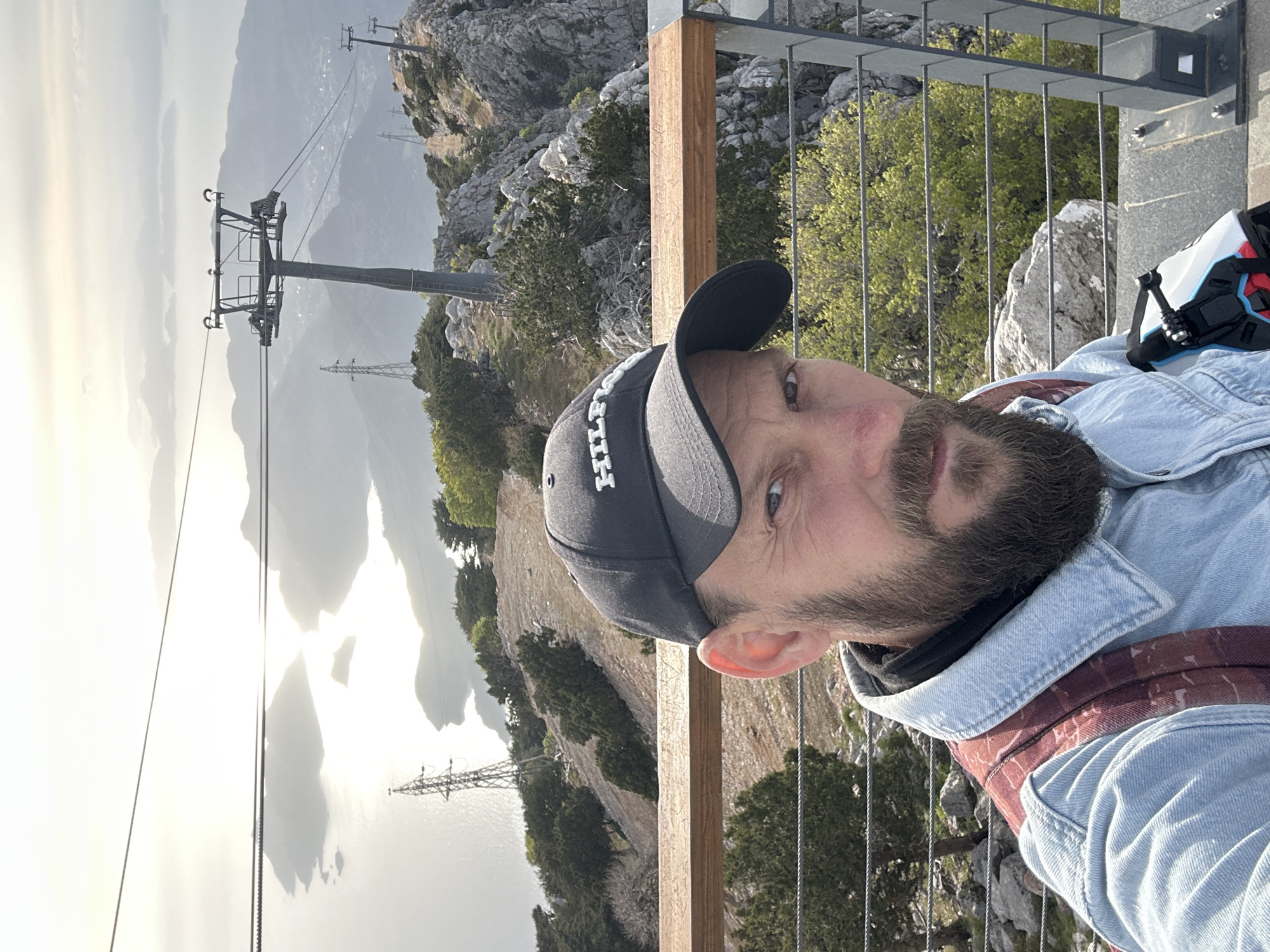 The image shows a man in the foreground wearing a baseball cap, a black shirt, and a denim jacket with a red backpack strap visible over his shoulder. Behind him, there is a mountain landscape. A ski lift can be seen on the left, stretching towards the distant mountains, partially obscured by clouds. The man is standing on a platform with a metal railing and wooden posts. The scenery suggests a high vantage point, likely a mountain peak or cliff overlooking a valley or body of water. The image appears to be taken at the Babadağ peak in Fethiye, Turkey, given the presence of the ski lift system that is unique to that mountain.