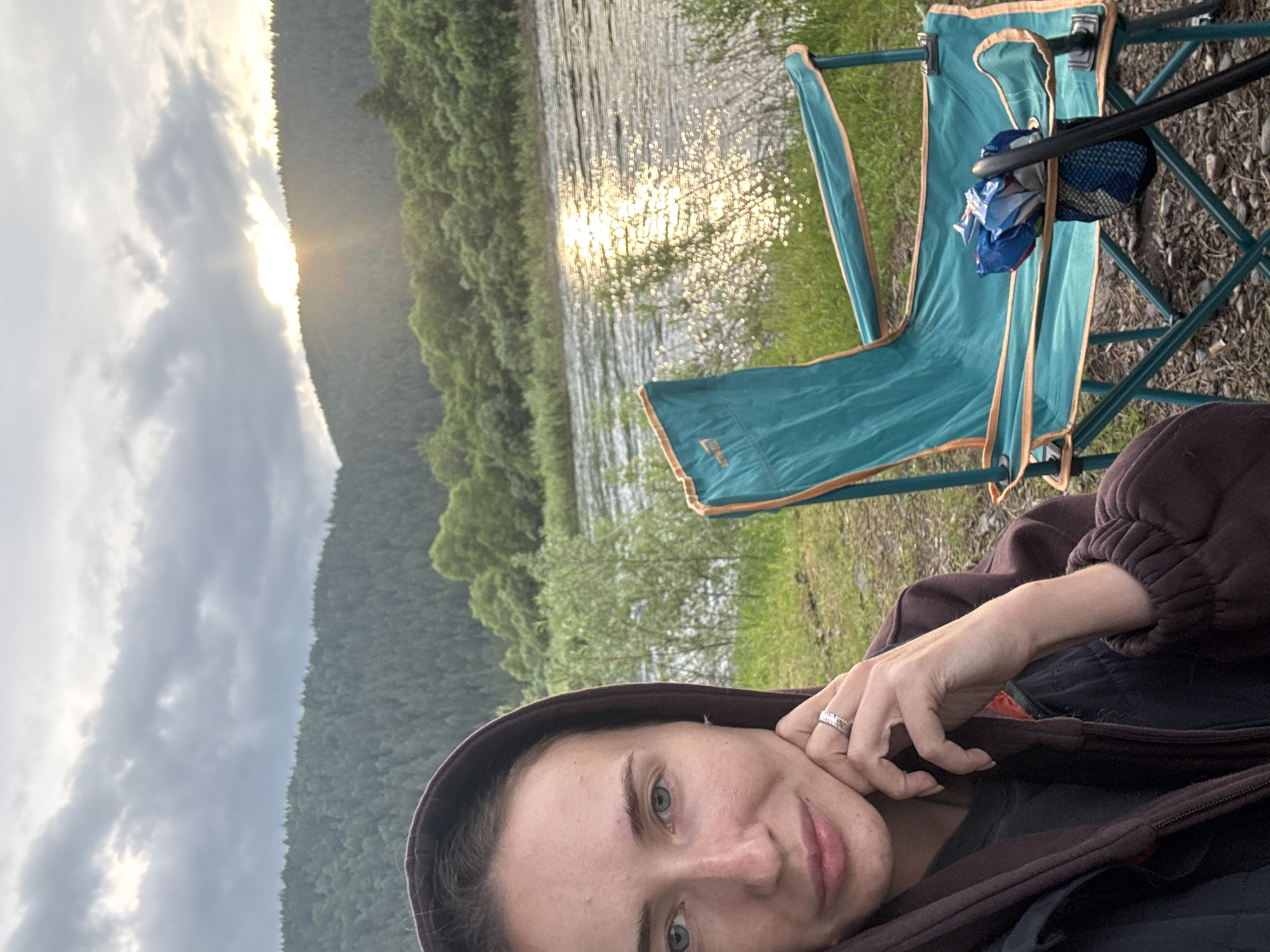 A young woman with light skin and blue eyes is pictured in a selfie. She is wearing a dark purple hooded jacket and has her hand on her chin, with a ring on her finger. Behind her, a teal camping chair with orange trim is visible, containing a bag of chips and a black mesh bag attached to its side. The background features a river with sunlight glinting off the water, a grassy embankment, and a densely forested hill under a cloudy sky with the sun peeking through. The scene appears to be in a natural, outdoor setting, likely a campsite by a river. The location is not the city of Narva, Russia; Narva is in Estonia.