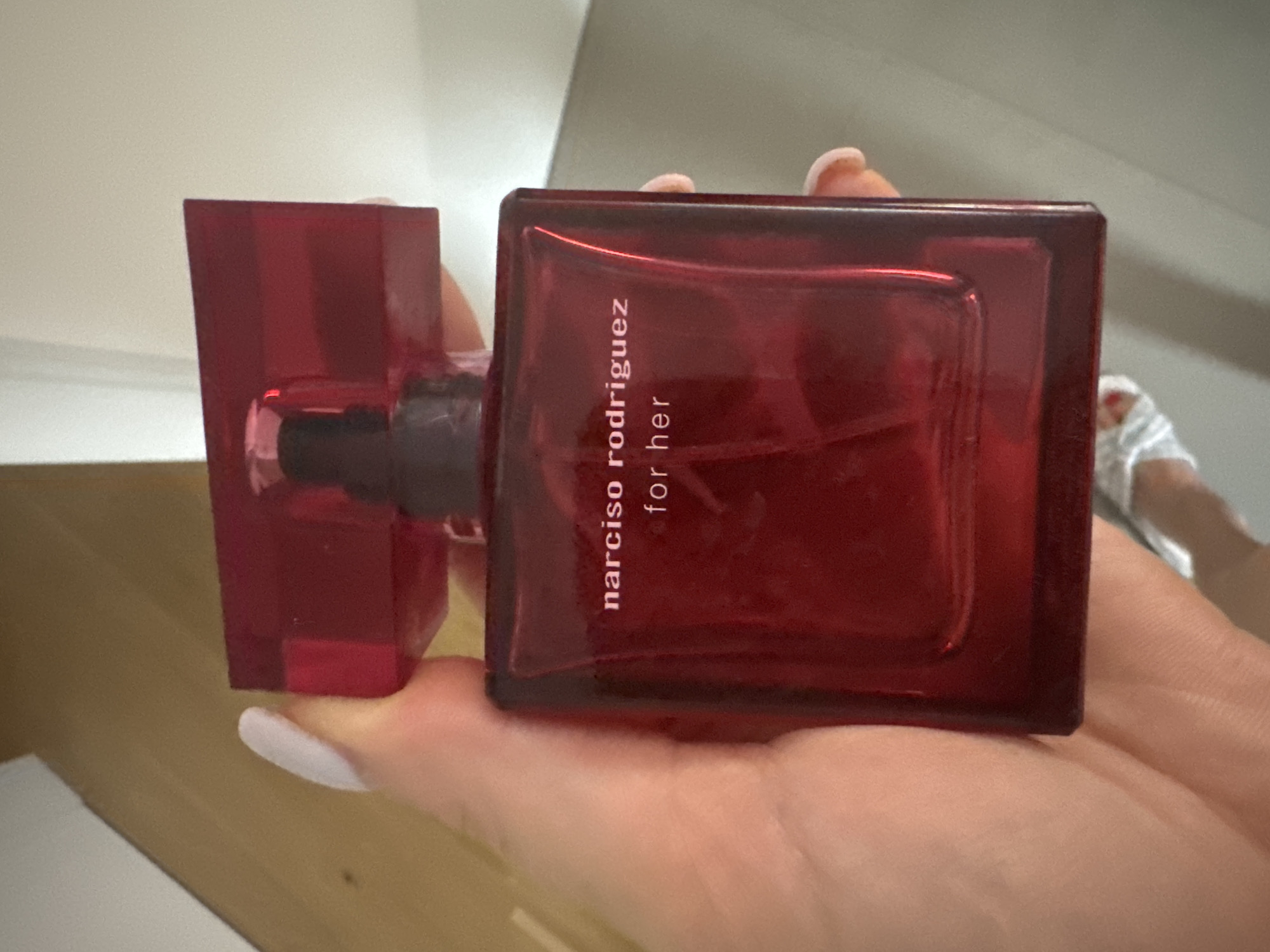 A person's hand, with manicured fingernails, is holding a red glass perfume bottle. The bottle has a dark cap and white text that reads "narciso rodriguez for her". The background appears to be an indoor setting with a wall and a floor. There are no visible landmarks or features that can definitively place this image in Murcia, Spain.