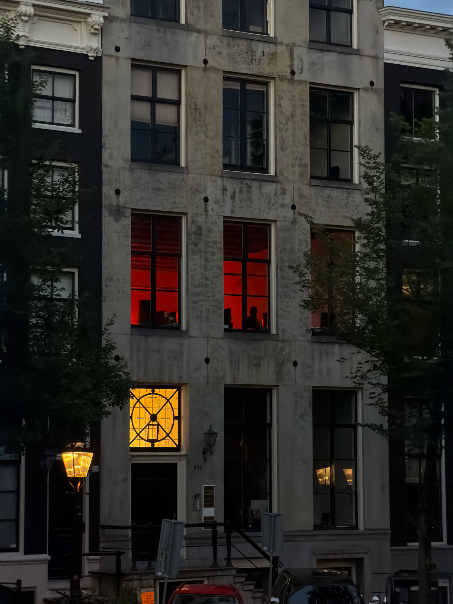 The image depicts the facade of a tall, narrow building in Amsterdam, Netherlands. The building appears to be a row house, with a darker facade on the left and a lighter, weathered concrete facade on the right. Several windows are visible on the building. Two upper-story windows on the right side of the concrete facade are illuminated with a vibrant red light, suggesting interior lighting. Below these, another window is illuminated with a warm yellow light and features a distinct circular pattern with geometric divisions, resembling a stylized clock or compass rose. A traditional Amsterdam-style street lamp, also lit with a warm yellow glow, is positioned in front of the lower part of the building on the left. Trees with dark green foliage partially obscure the facade on both sides of the image. The sky above is a dusky blue, indicating either early morning or late evening. A partial view of cars and street furniture is visible at the bottom of the frame. The building's architecture is typical of Amsterdam's canal-side housing.