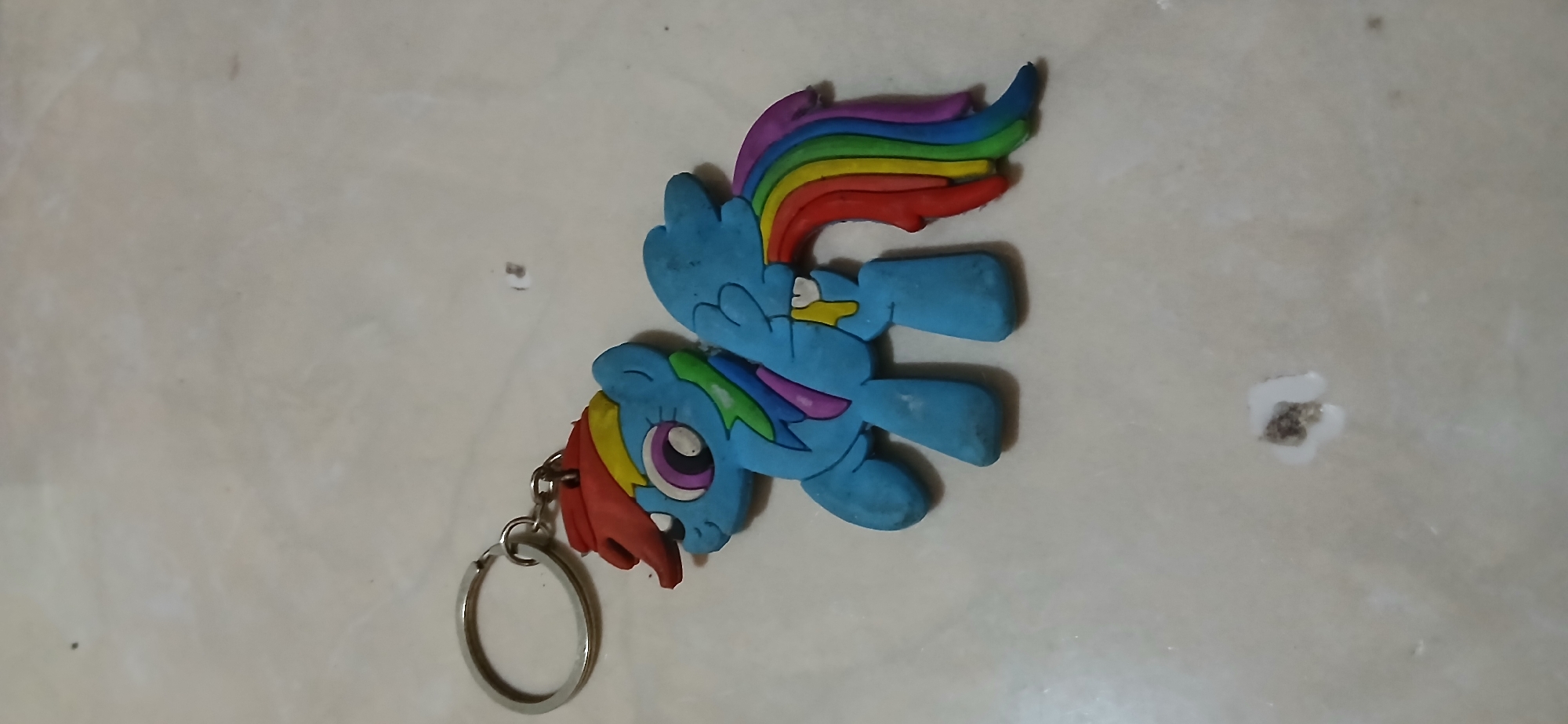 A keychain featuring a blue cartoon pony with a rainbow-colored mane and tail is resting on a light-colored, textured surface that appears to be a marble countertop. The pony is in a walking pose, with its head turned slightly to the left, revealing large, purple-and-white eyes. Its mane is a mix of red and yellow, and its tail is a vibrant cascade of red, orange, yellow, green, blue, and purple. The pony has a small yellow detail on its chest and two white teeth visible. The metal keychain ring is attached to the pony's head by a small chain. The surface beneath the keychain shows minor dirt and a few small, dark specks, along with a small white substance with a dark center. The lighting suggests an indoor environment. This item was likely photographed in Samarinda, Indonesia, a city known for its artisan crafts and the common use of decorative items like this keychain in daily life.