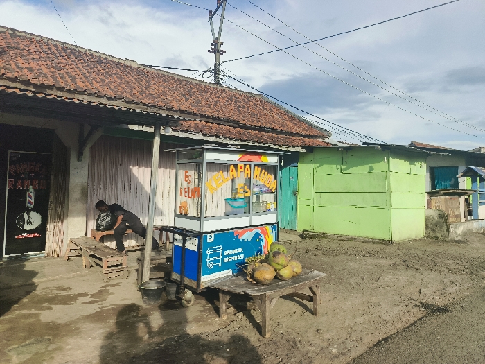 The image shows a street scene in Cikalong, Indonesia (latitude: -6.759, longitude: 107.447). A man in a black shirt and pants is standing by a wooden table, bending over as if placing something down. The table is in front of a small storefront with a tiled roof and a black door with the word "RAMBUT" in white letters. The storefront is part of a larger building with a tile roof and a wooden overhang. To the right of the storefront, there is a small food cart with a blue and white sign that says "ES KELAPA MUDA". The cart is stocked with coconuts and a small cooler. There are wooden benches on the ground in front of the storefront and the food cart. A second, green-painted building with a tile roof is located to the right of the food cart. There are power lines above the scene. The ground in the image is dirt and gravel. The weather is cloudy.