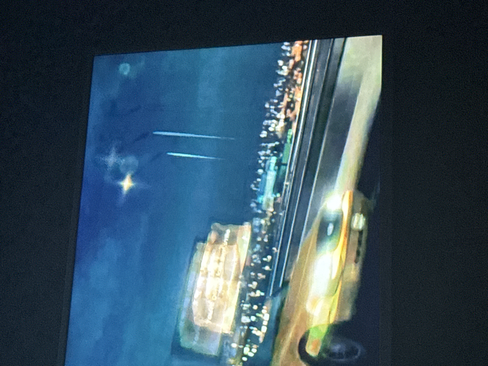 The image appears to be a photograph of a screen displaying a night scene, likely an image or video of a cityscape. A body of water is on the left with a prominent star visible in the sky. In the center is a building with many lights. On the right side is a road with cars moving on it. Given the question, it is most likely an image of Mytishchi, Russia, at night, but specific locations cannot be determined.