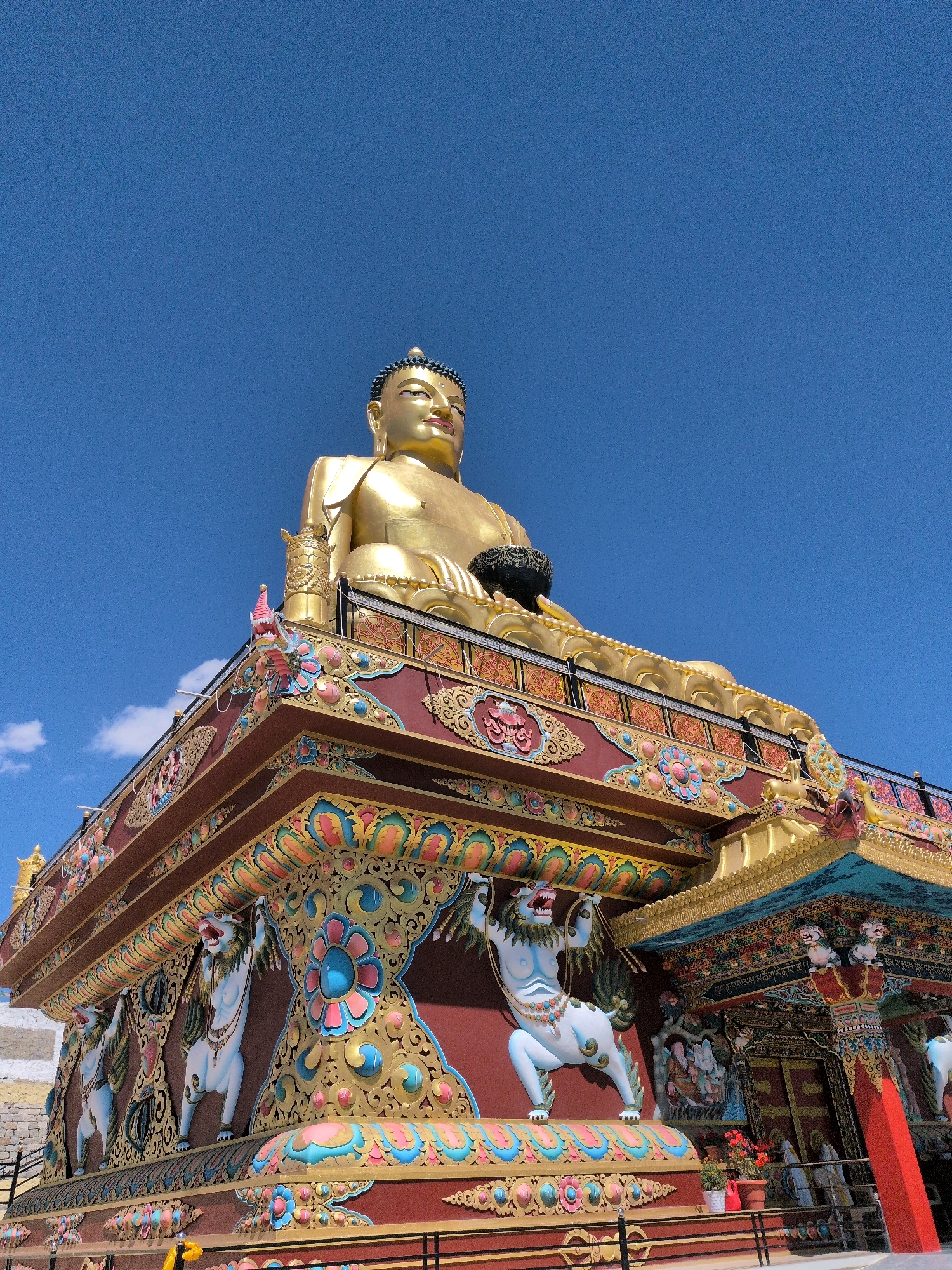This is a low-angle, outdoor, full-shot photograph of a large, golden Buddha statue situated atop a vibrantly decorated structure. The Buddha is seated, with its legs crossed and hands in a meditative pose, holding a dark bowl. The statue is gilded and detailed, with intricate facial features and a blue, crown-like headdress.
The structure supporting the Buddha is a multi-tiered edifice adorned with colorful Buddhist motifs. The primary color of the structure's facade is a deep maroon, contrasted with intricate patterns in gold, blue, pink, and teal. Notable decorative elements include stylized lion-like figures supporting the roof, ornate floral designs, and what appear to be depictions of celestial beings or deities within recessed panels. The roof is capped with golden embellishments.
The sky above is a clear, bright blue with a few wispy white clouds visible on the left side. The environment appears to be arid, with hints of a stone or earthen wall in the background on the left. The overall impression is one of grandeur and spiritual significance. This statue is a prominent landmark in the city of Leh, Ladakh, India, known as the Maitreya Buddha Temple or Leh Buddha.