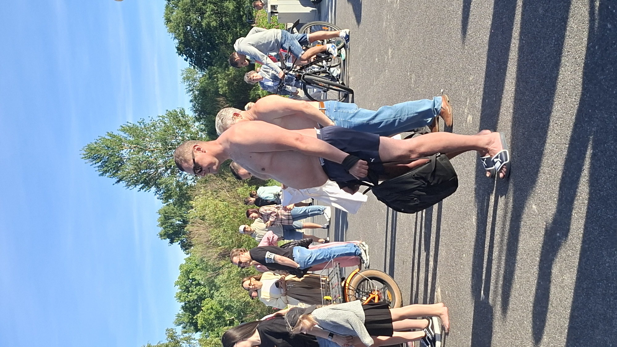 The image shows a group of people standing in a line outdoors on a sunny day. The scene appears to be a street or pavement next to a grassy area with trees. The people are facing away from the camera and are dressed in casual clothing, with several men shirtless. Some individuals are standing with their hands behind their backs. A bicycle with a prominent orange frame is visible towards the right side of the image, positioned behind some of the people. The shadows cast by the people and an unseen railing indicate that the sun is high in the sky, suggesting it is midday. The sky is a clear blue. The overall atmosphere seems relaxed and casual. There is no visible text in the image.