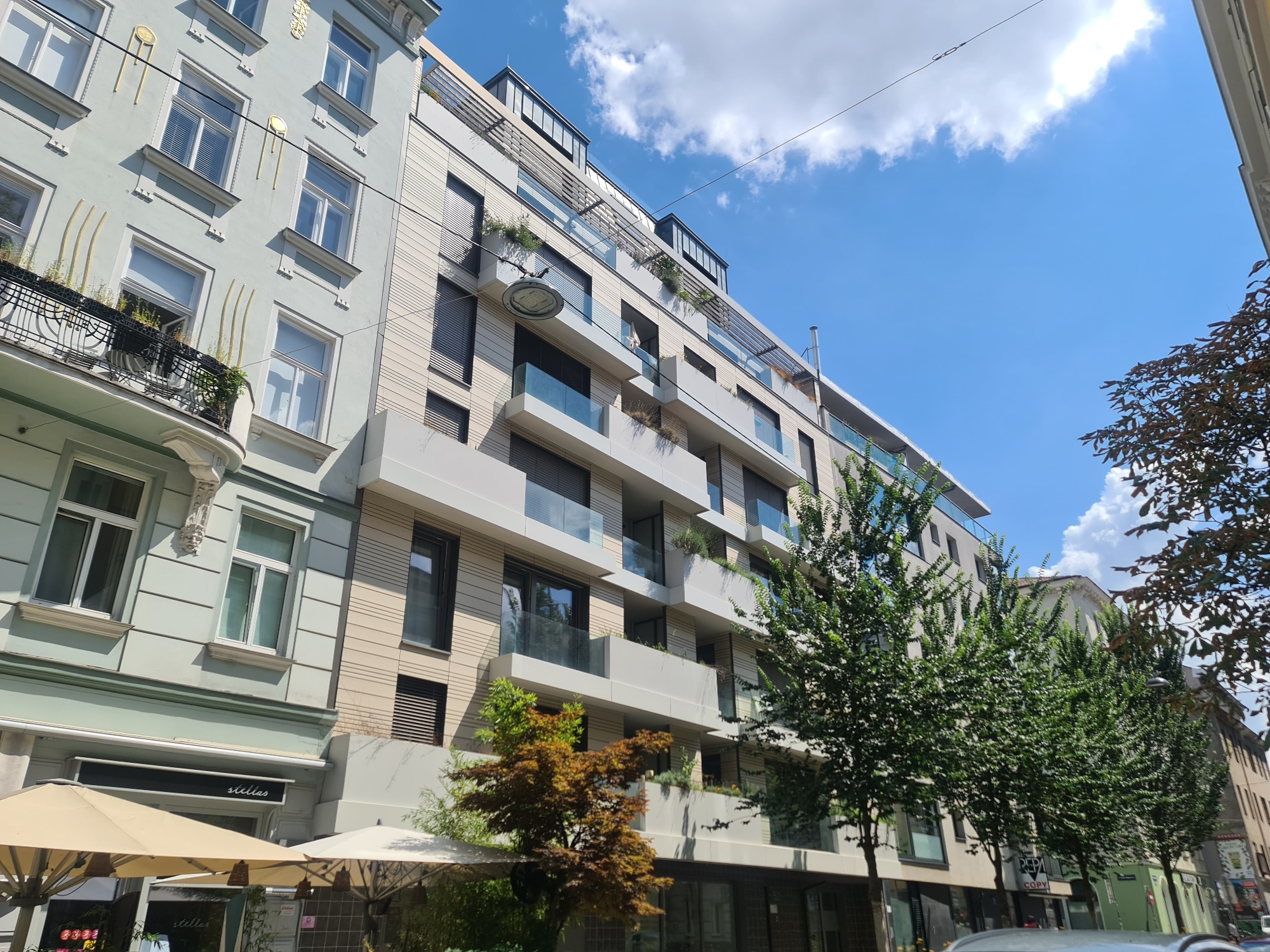 The image depicts a street scene in Vienna, Austria, characterized by two adjacent buildings of contrasting architectural styles. The building on the left is older, with a pale green facade, classical decorative elements, and balconies adorned with wrought iron. The building on the right is a modern apartment complex with a neutral color palette, featuring multiple floors with balconies, glass railings, and numerous windows. Trees line the sidewalk in front of the modern building, their green foliage contrasting with the urban architecture. The sky above is a vibrant blue with scattered white clouds, indicating a sunny day. Umbrellas suggesting an outdoor seating area for a cafe or restaurant are visible in the foreground, in front of the older building. The scene captures a slice of urban life in Vienna, showcasing the coexistence of historical and contemporary architecture.