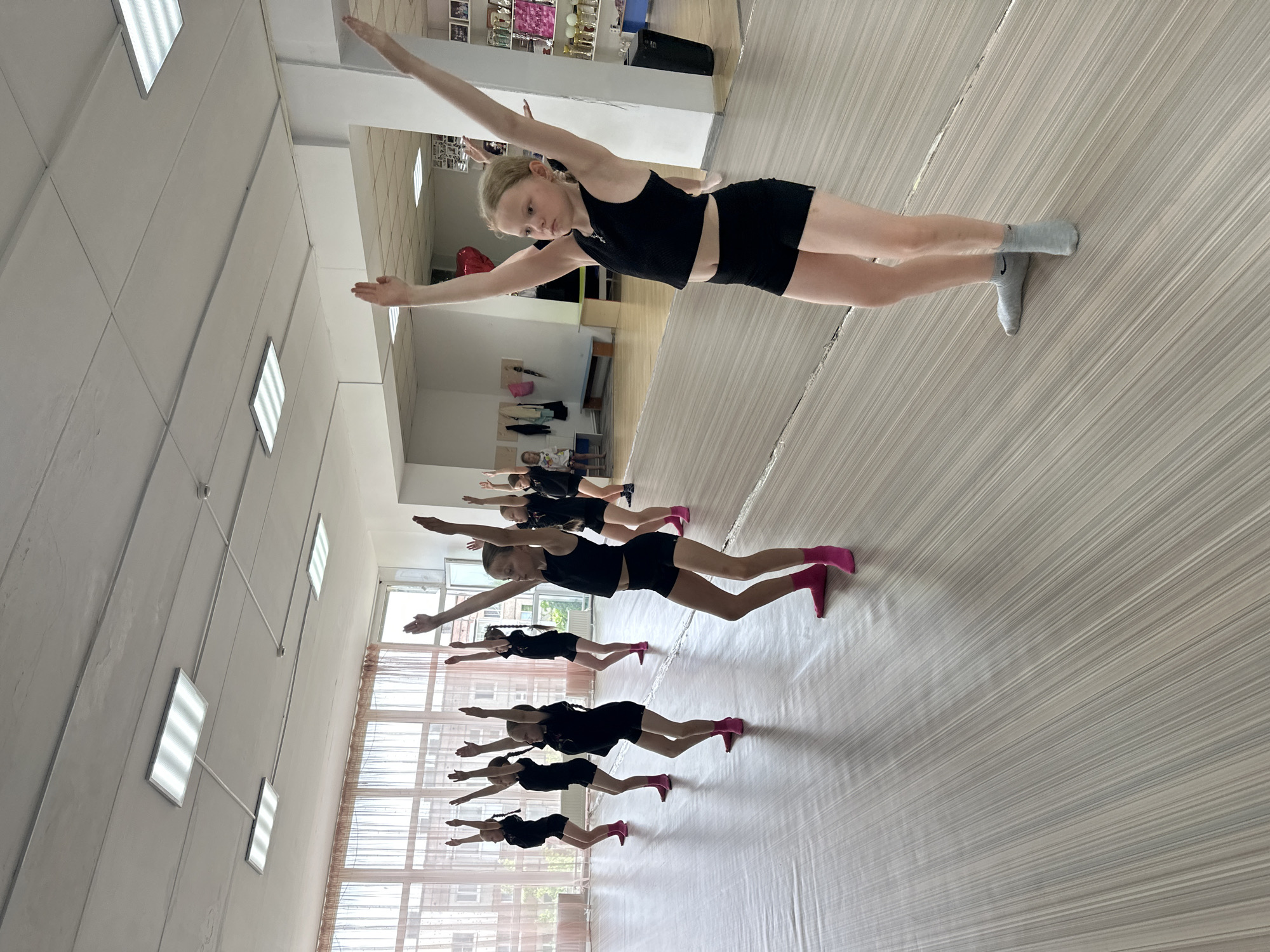 The image shows a group of young girls, possibly ballet or dance students, in a dance studio in Votkinsk, Russia. They are arranged in a line and performing a balancing exercise, with arms extended and one leg raised. They are wearing black shorts and tops, with some wearing pink ballet shoes. The studio has a light-colored, striped floor, light walls, and fluorescent ceiling lights. A mirror is present on the front wall of the room, and windows can be seen behind the dancers, indicating the presence of a window. The studio also has a storage area or room in the back.