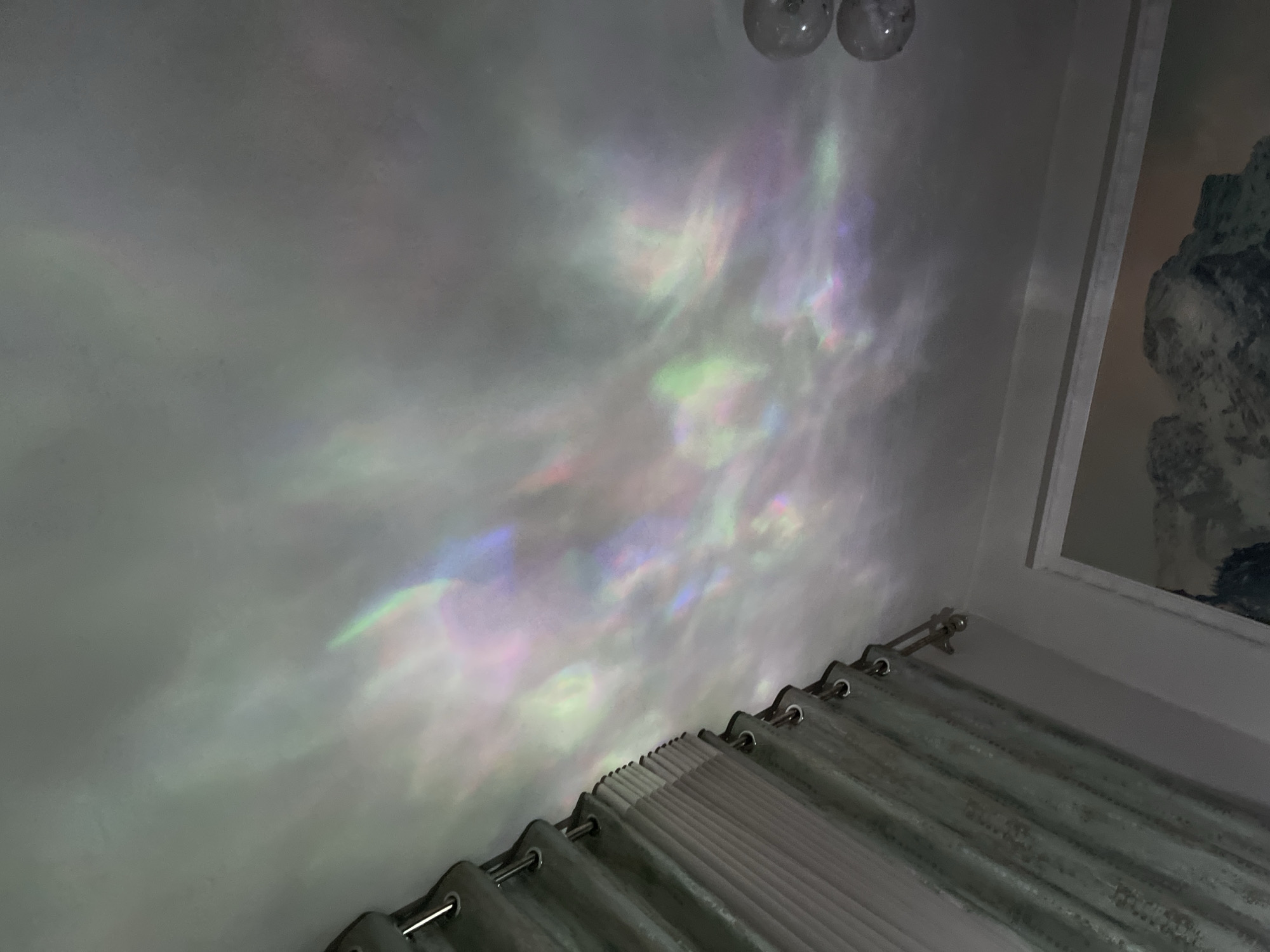 The image shows a ceiling illuminated by colorful, nebulous light patterns, resembling the aurora borealis. To the left, a section of a pale green, textured curtain with grommet holes is visible, hanging from a curtain rod. In the upper right corner, two spherical objects, possibly lights or decorative elements, are partially visible. The overall impression is of an indoor space with ambient lighting. The location is identified as Bishkek City, Kyrgyzstan.